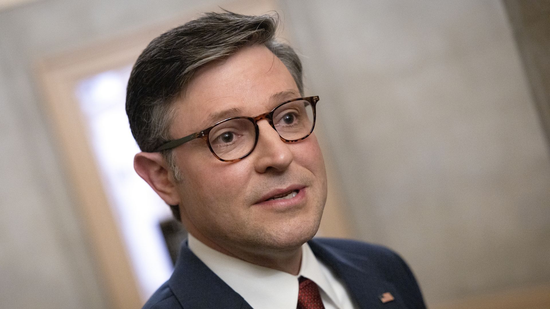 House Speaker Mike Johnson, wearing a blue suit and standing in a sandstone corridor.