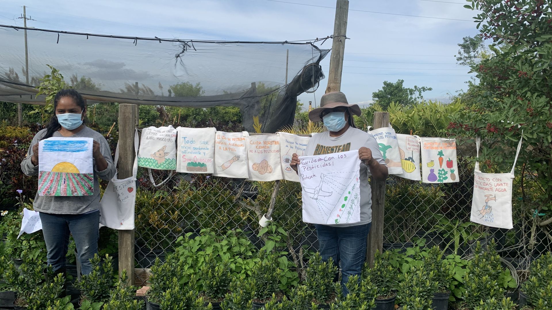 Two women stand in a field while holding up custom-made tote bags used to protest farmworkers' exposure to pesticides. 