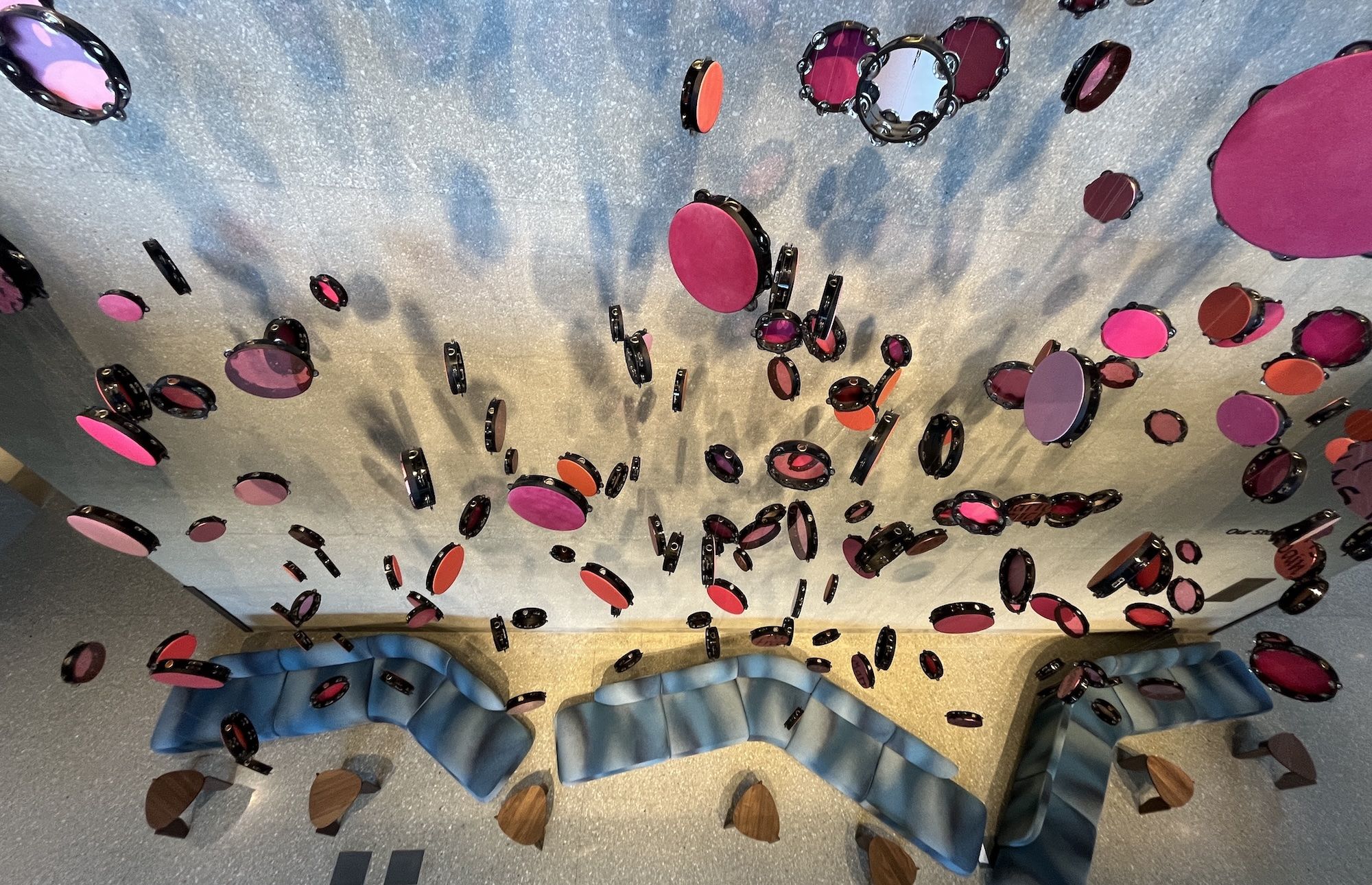 Floating circular discs in pink, magenta, and orange with black rims drift above a blue backdrop ceiling, creating a colorful installation and casting soft shadows.