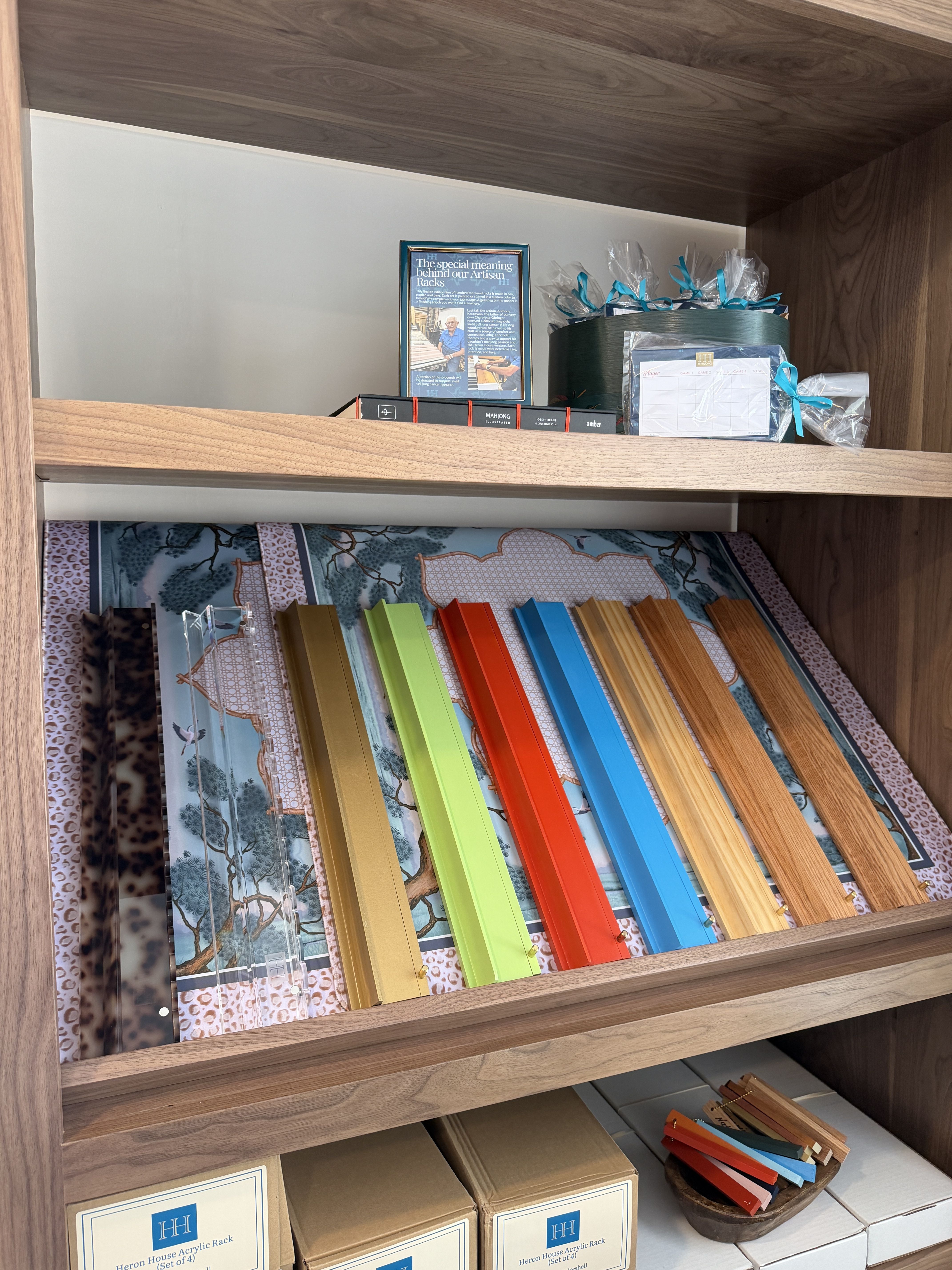 Wooden shelf filled with craft items: a framed article, a gift box with teal ribbons, and a rainbow row of acrylic color strips; lower boxes read "Heron House Acrylic Rack".
