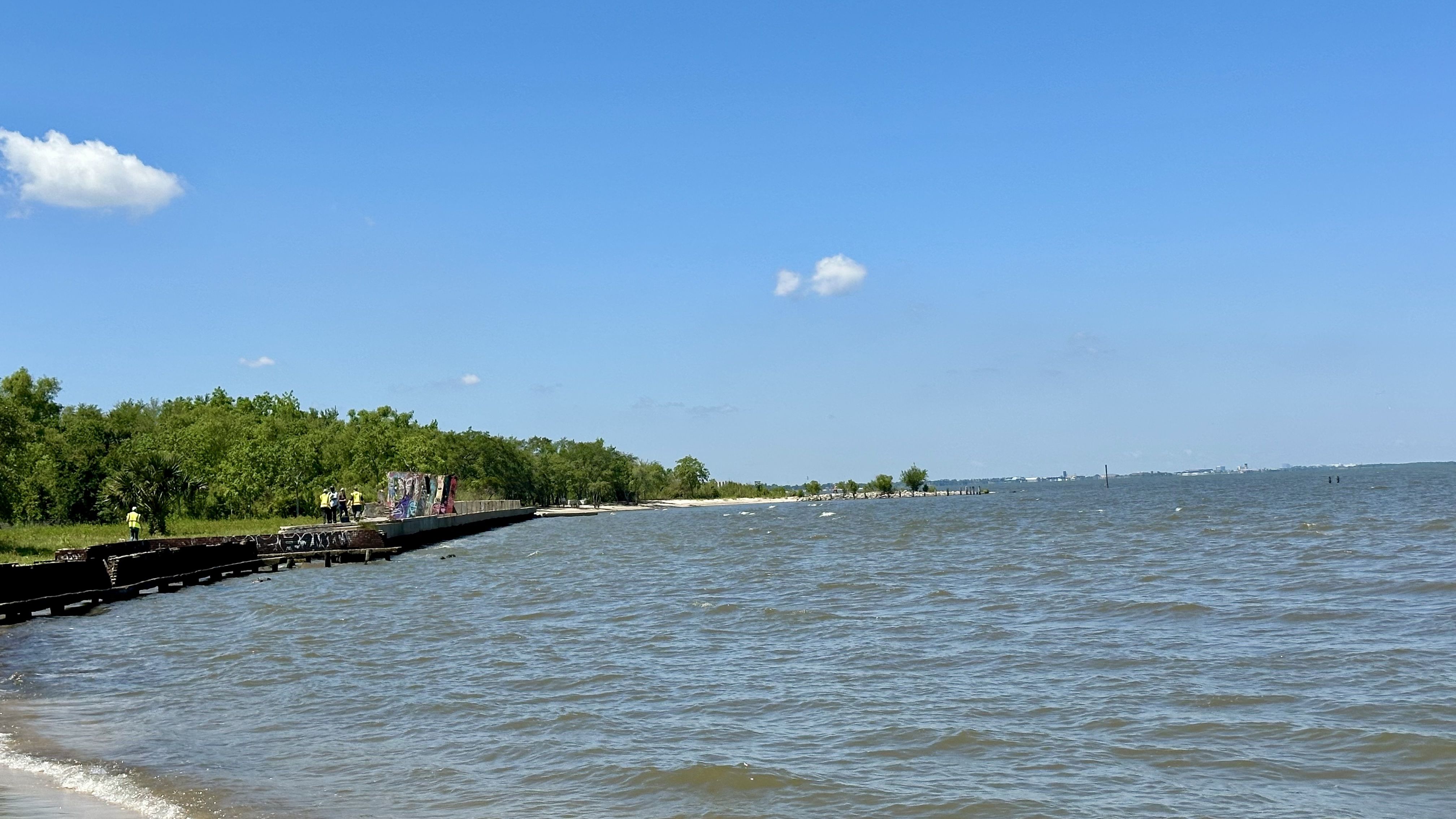 A blue sky with a few clouds over a large body of water, a green tree-lined shore, and a breakwater or seawall with graffiti and people wearing yellow vests.