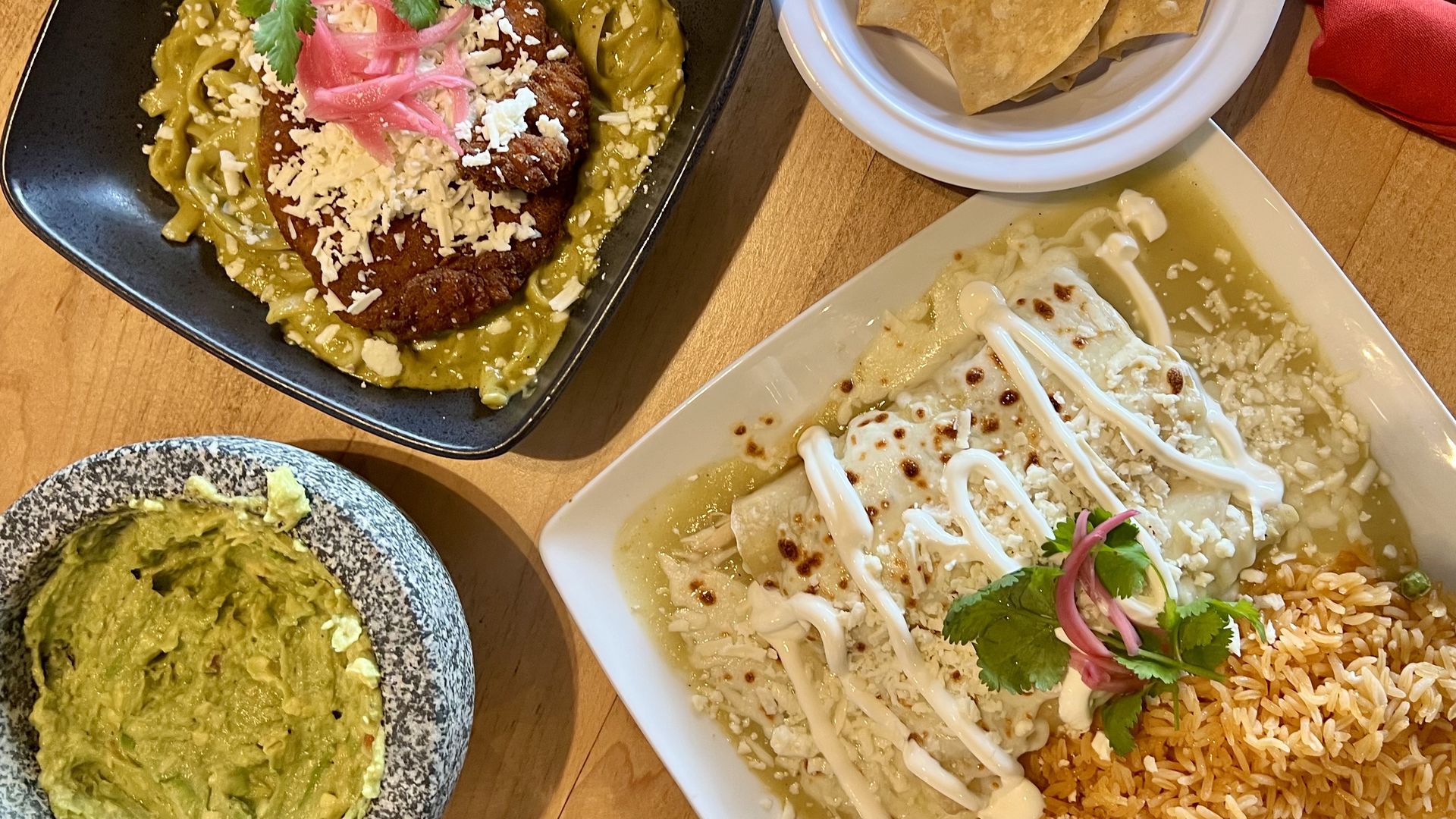 Mexican dishes including green sauce enchiladas with sour cream and cheese, a plate of refried black beans topped with cheese and pickled onions, a bowl of guacamole, and a side of tortilla chips on a wood table.