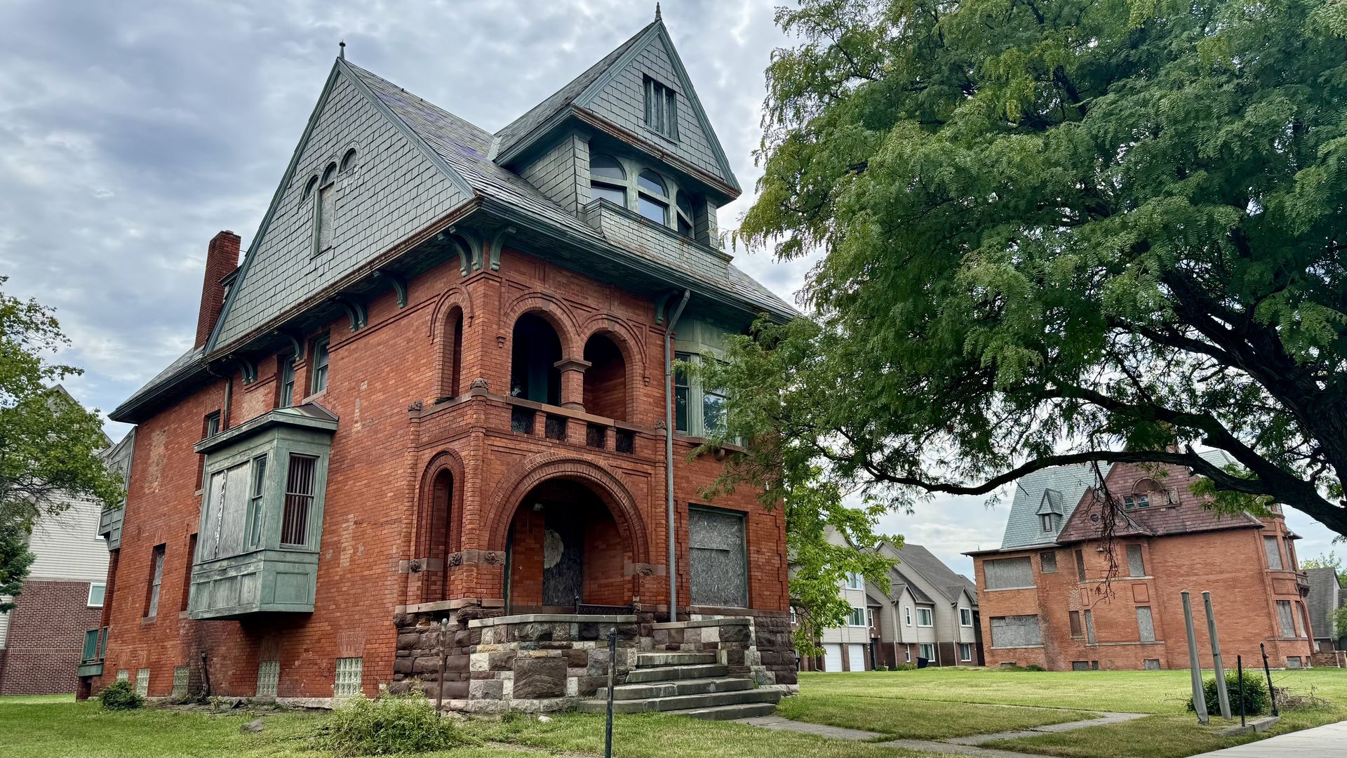 Red brick Victorian house with arched brick porch, gray slate roof and tower, set on stone foundation; some windows boarded up.