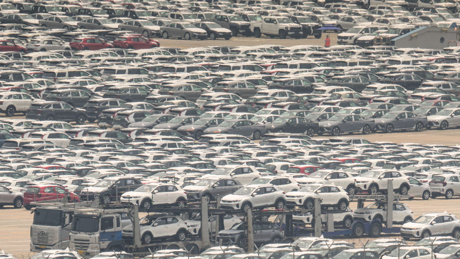 Large lot filled with hundreds of white and some dark-colored new cars closely parked in rows, with car carriers loaded with vehicles in the foreground and light haze overhead.