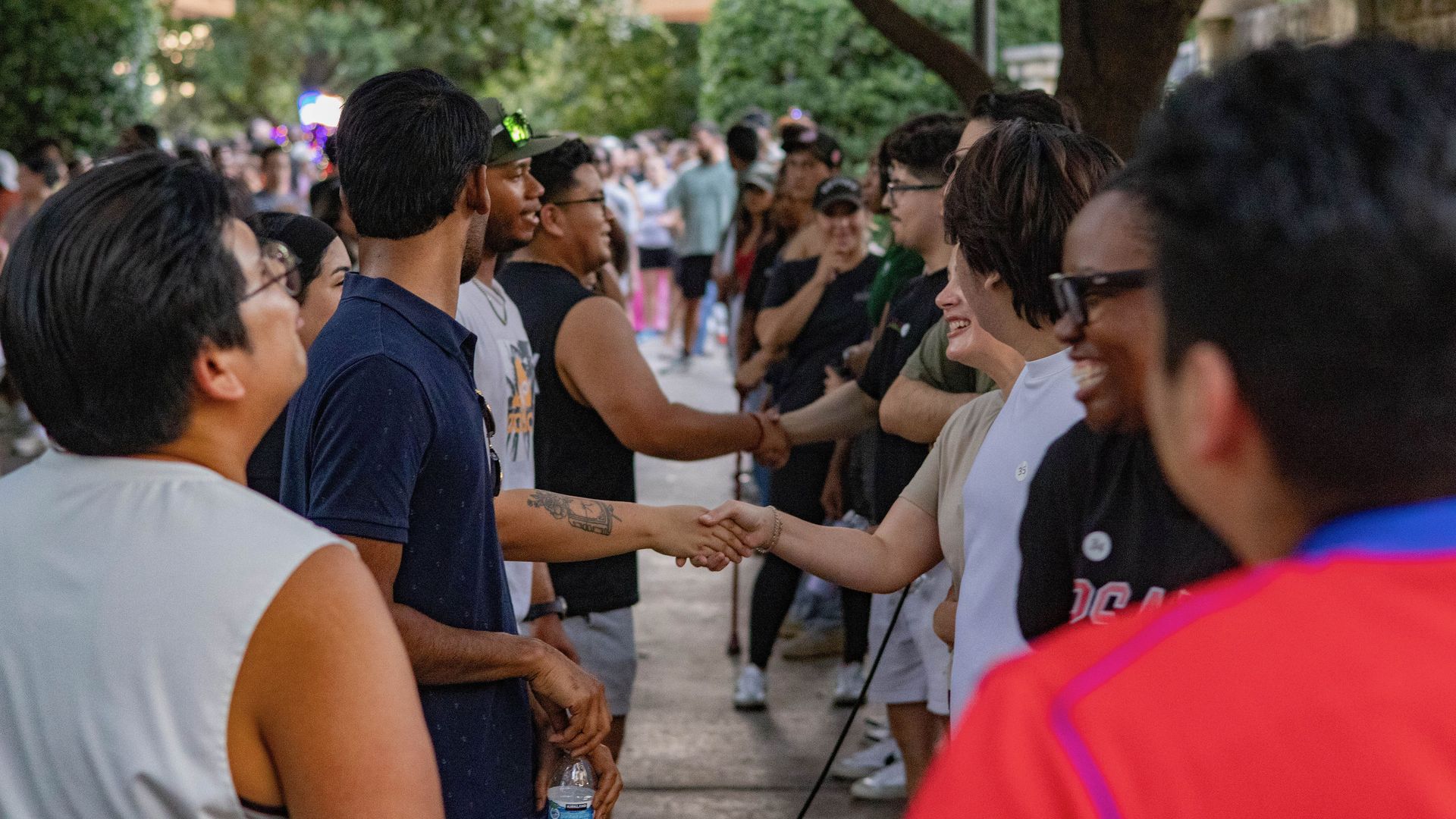 Outdoor park scene with a long line of people shaking hands face-to-face; foreground shows a man in a dark blue shirt shaking hands with someone in white, trees in the background.