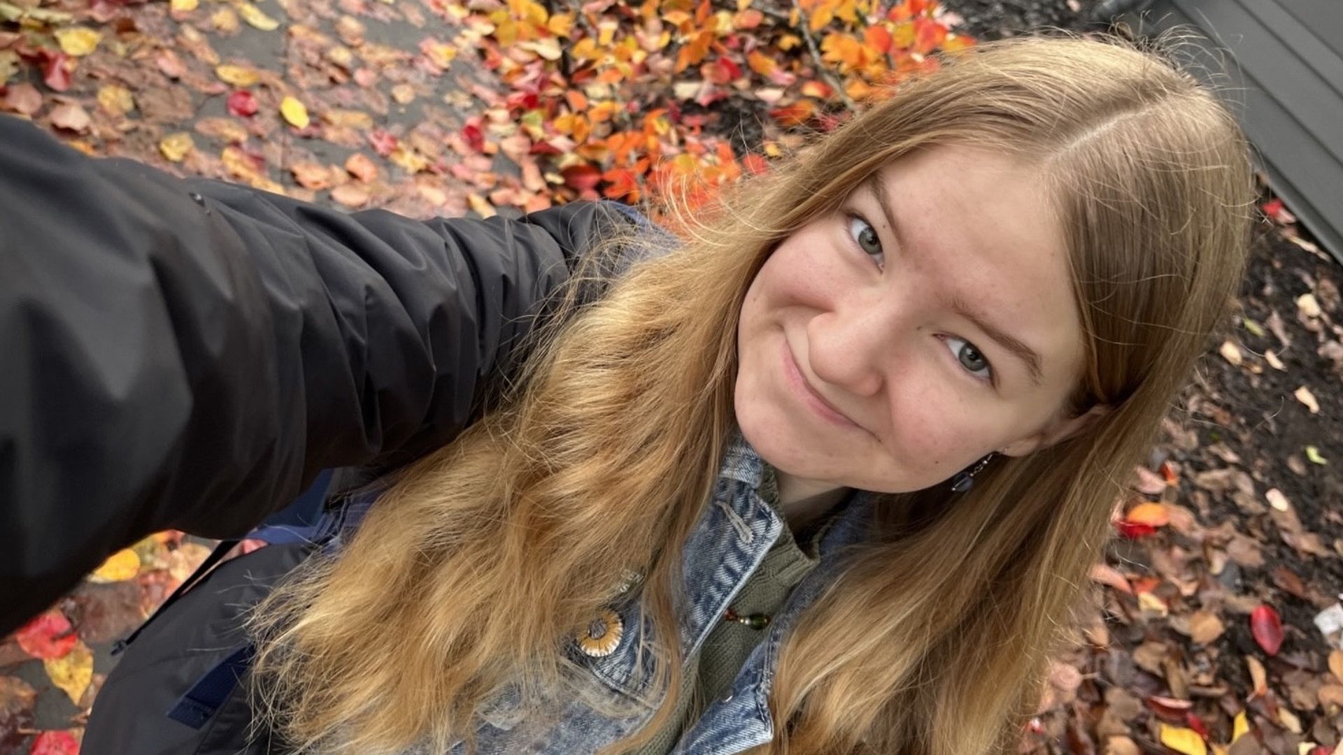 A girl with blondish hair past her shoulders smiles in a selfie. The sidewalk behind is wet and covered in small orange and red leaves.