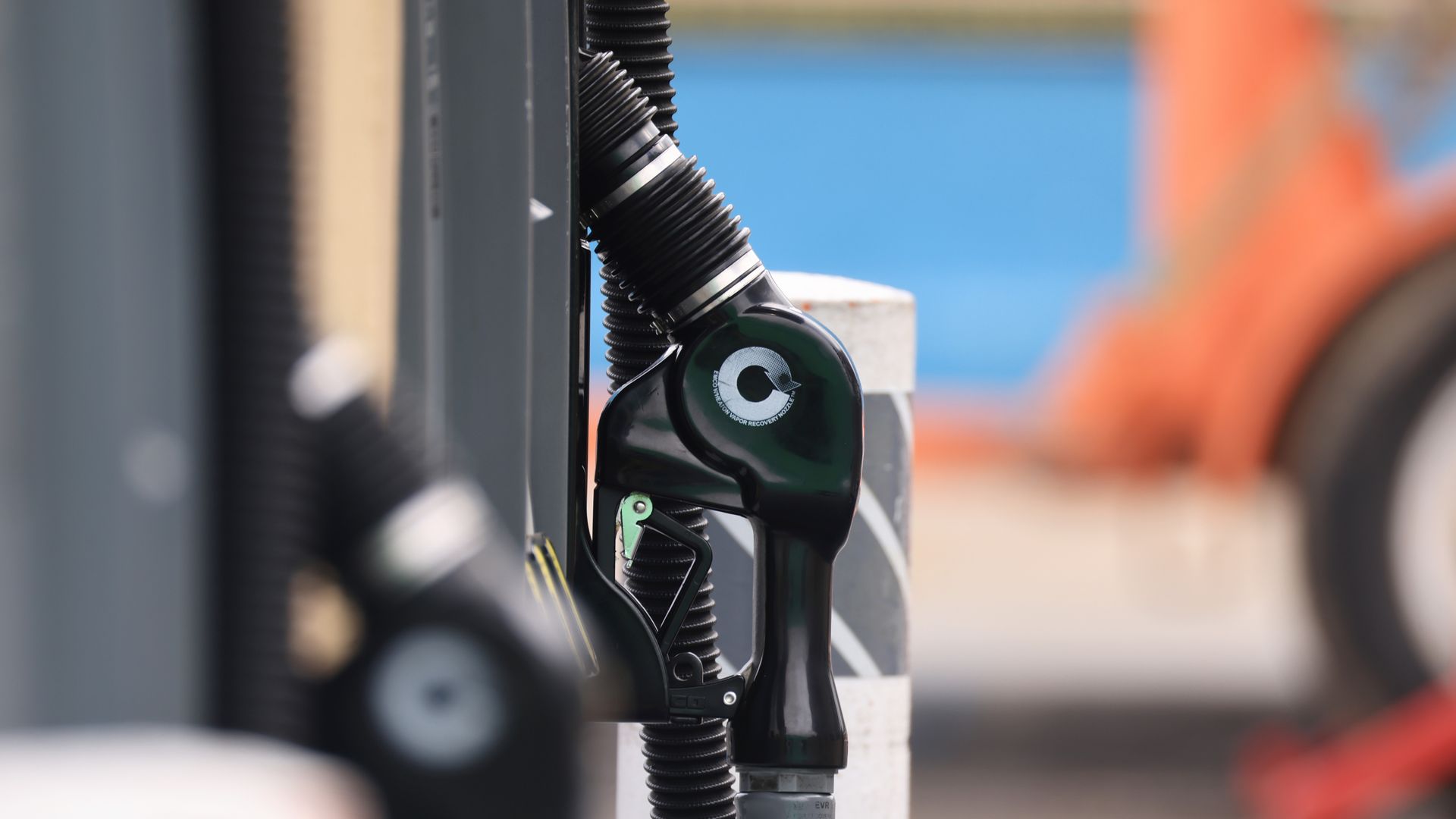 Close-up of a green gas pump nozzle attached to a black ribbed hose, mounted at a fueling pump with a blurred blue background.