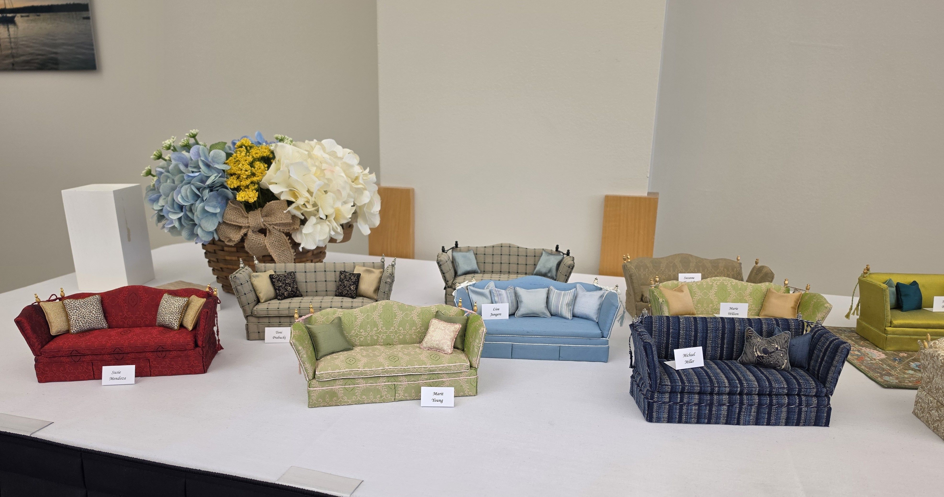 Display of miniature upholstered sofas in red, green, blue, and beige with cushions, arranged on a white table; a wicker basket of blue and white hydrangeas sits behind.