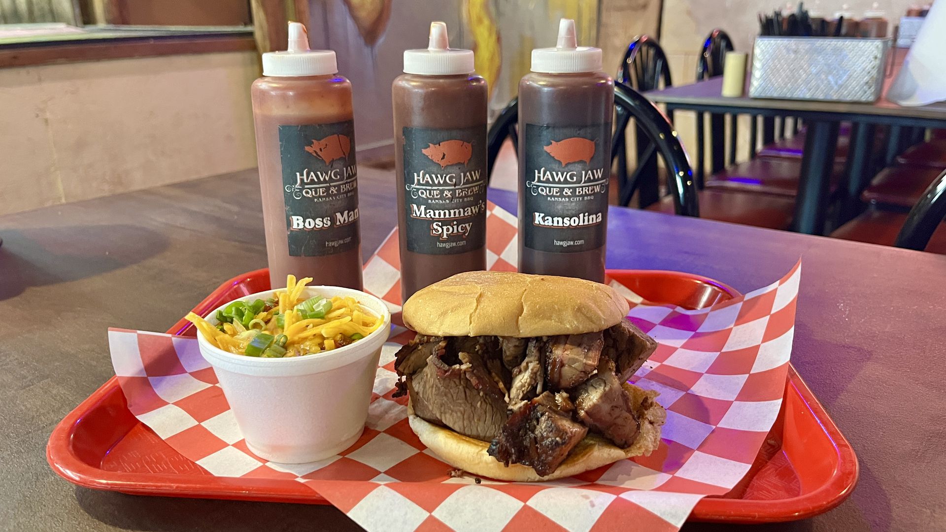 Barbecue sandwich with smoked meat on a bun, a side of cheesy baked beans topped with green onions in a white cup, and three BBQ sauce bottles labeled Boss Man, Mammaw's Spicy, and Kansolina on a red tray.