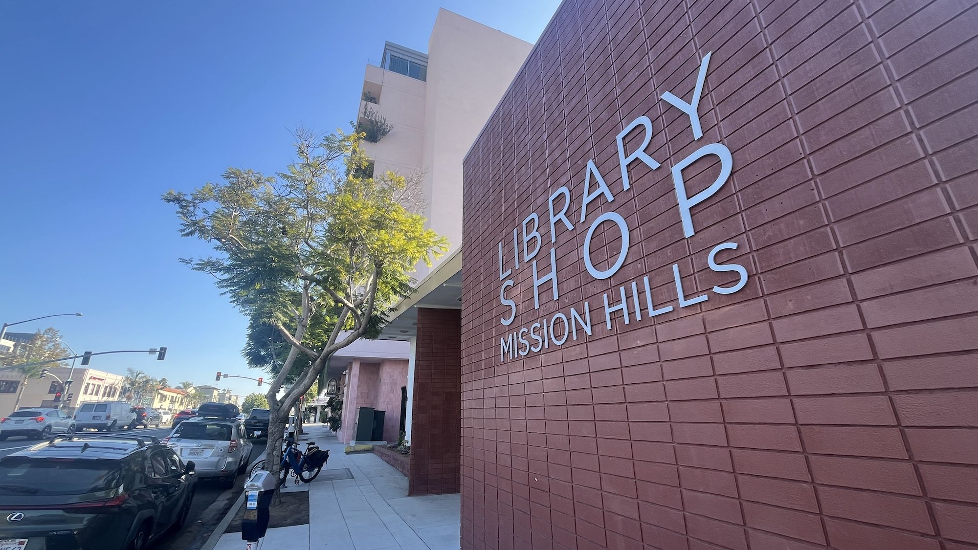 Red brick building with silver letters reading "Library Shop Mission Hills" beside a tree-lined sidewalk and parked cars under a clear blue sky.