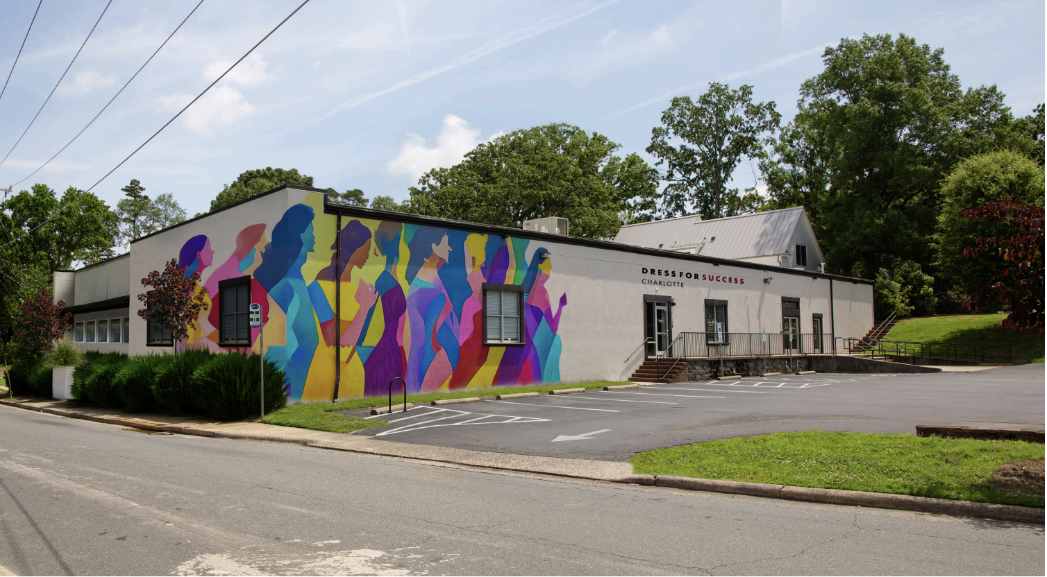 Building with a colorful mural of women's silhouettes in vibrant colors on one side, labeled "Dress for Success Charlotte," surrounded by trees and an empty parking lot.