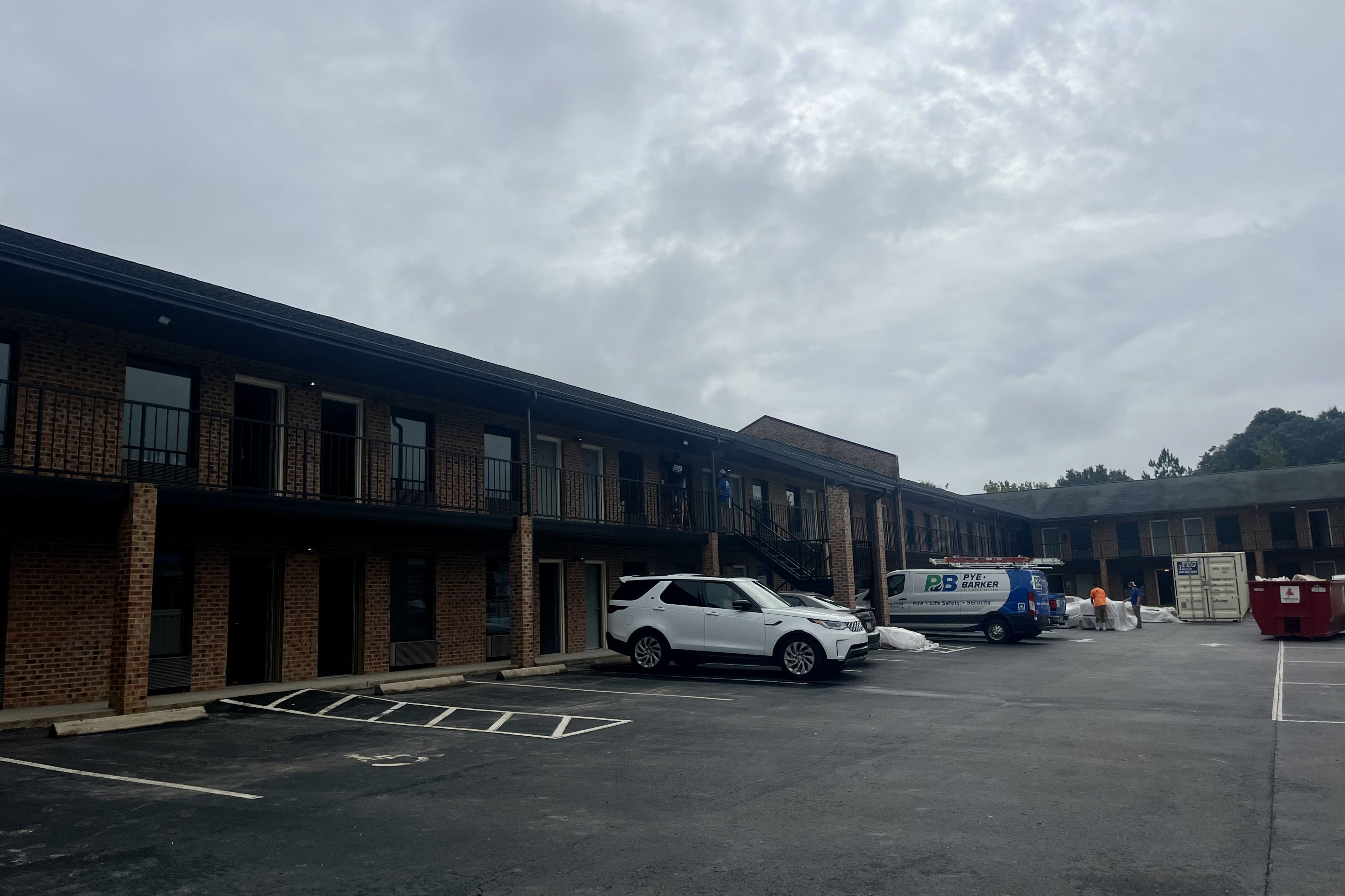Overcast sky above a two-story brick motel with black railings and exterior staircases, a white SUV and a van parked in the asphalt parking lot, workers near red dumpster and white containers.