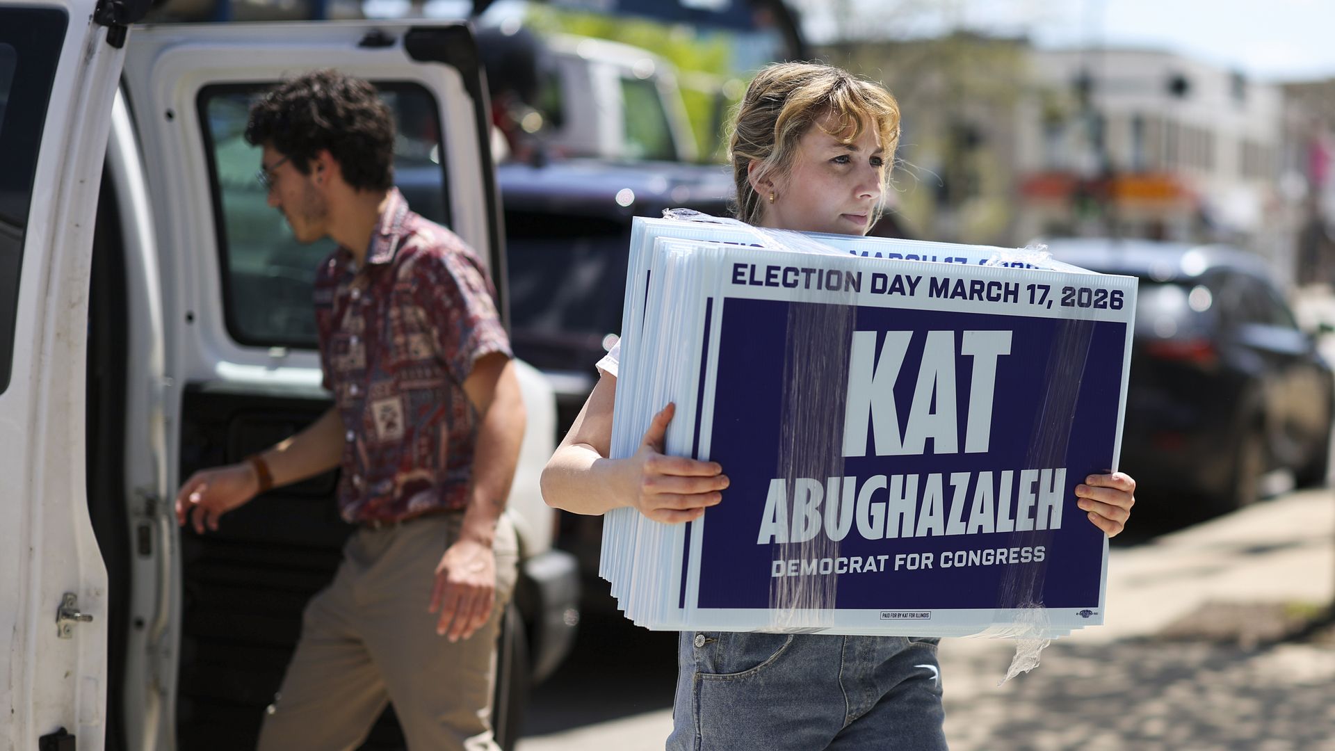 Kaat Abughazaleh, holding signs in front of a row of cars with a campaign worker reaching into a white van.