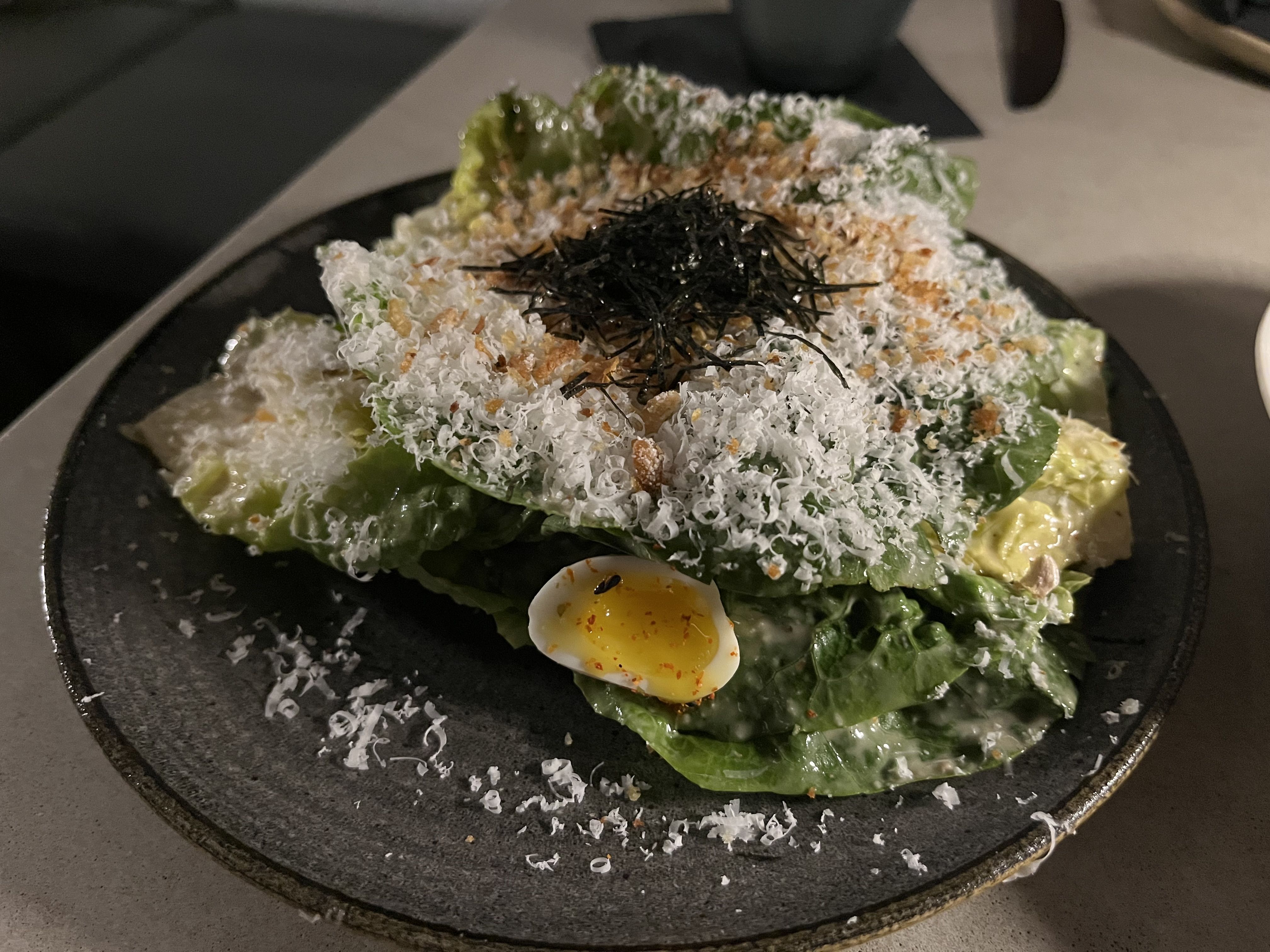 Salad with romaine lettuce, grated white cheese, fried garlic bits, shredded dark seaweed, and a halved soft-boiled quail egg on a dark plate.