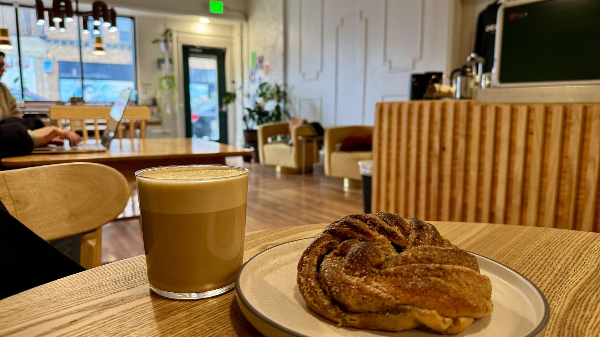 A café scene with a latte and a pastry on a wooden table, people working on laptops and cozy beige chairs in the background.