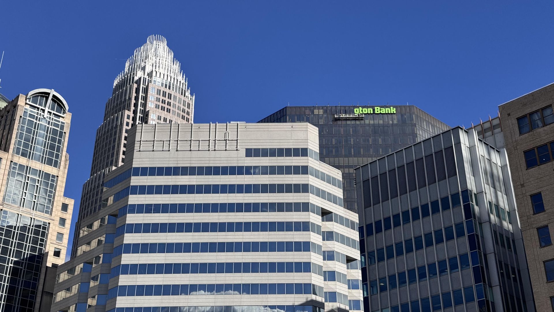 Clear blue sky over a cluster of modern glass office towers, including a tall white-fin skyscraper; a gray building in the foreground features a green Bank sign.