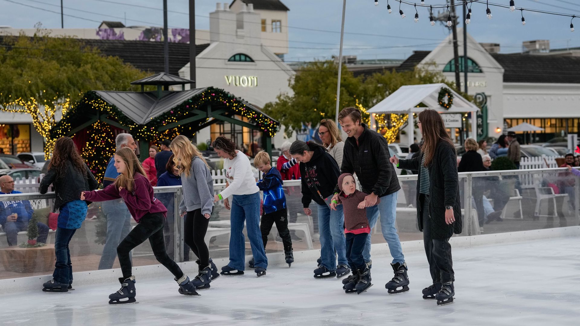 People of various ages ice skating outdoors in a decorated rink with string lights, holiday wreaths, and illuminated trees, near buildings and parked cars at dusk.