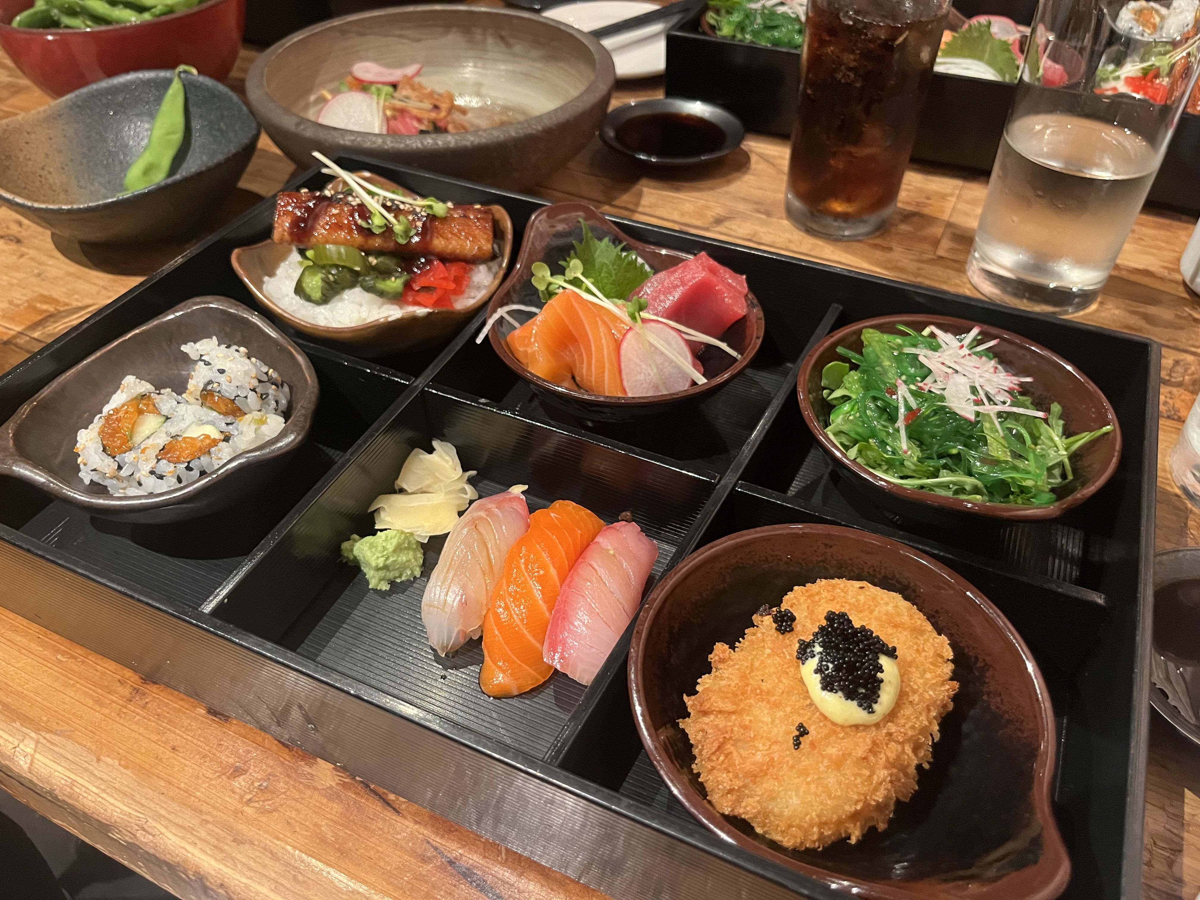 Top-down view of a Japanese bento spread: sushi and sashimi (salmon, tuna), eel on rice, greens, pickles, sesame rice, a crispy croquette with mayo and caviar, plus cola and water on a wooden table.
