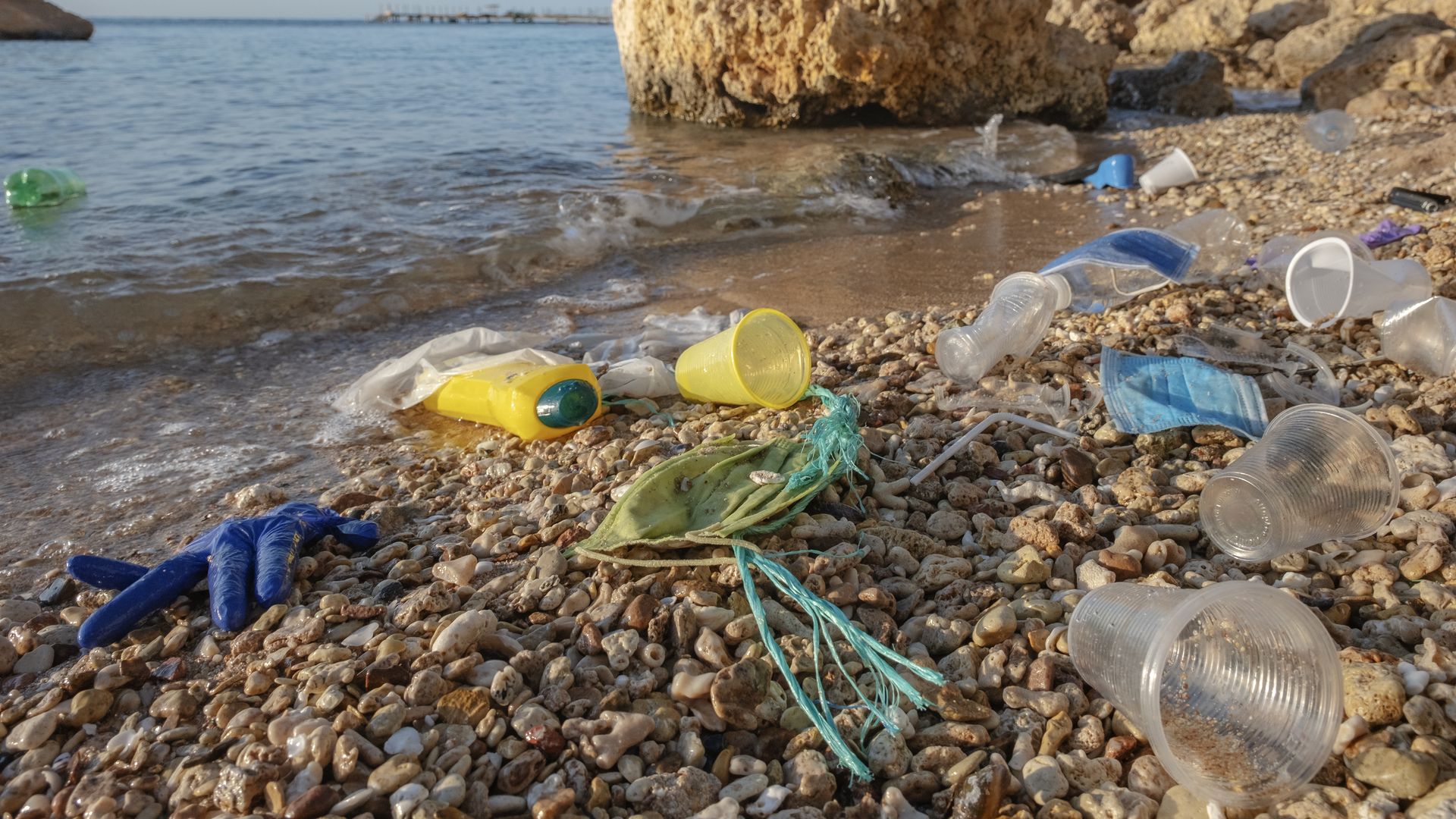 Photo of plastic waste, including a rubber glove, plastic cup and face mask, on a sea shore next to a body of water