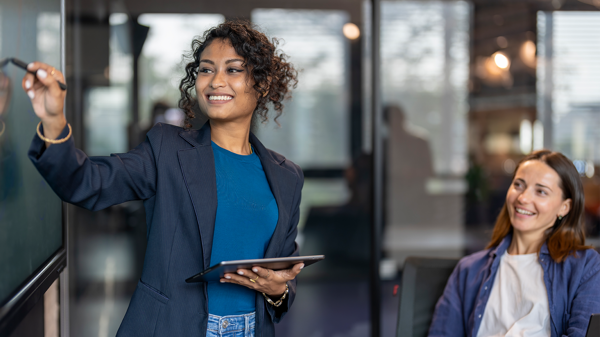 Smiling woman in a blue shirt and dark blazer writing on a board while holding a tablet, with another woman in a white shirt and blue jacket sitting and smiling in a modern office.