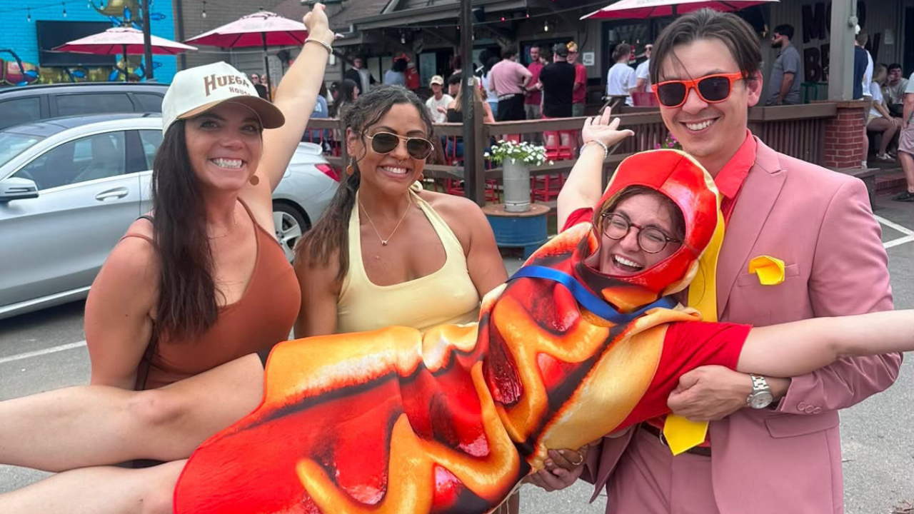 Four friends pose at a sunny outdoor festival: a man in a pink suit and orange sunglasses holds a person wrapped in a red-orange flame blanket; two women smile beside them.
