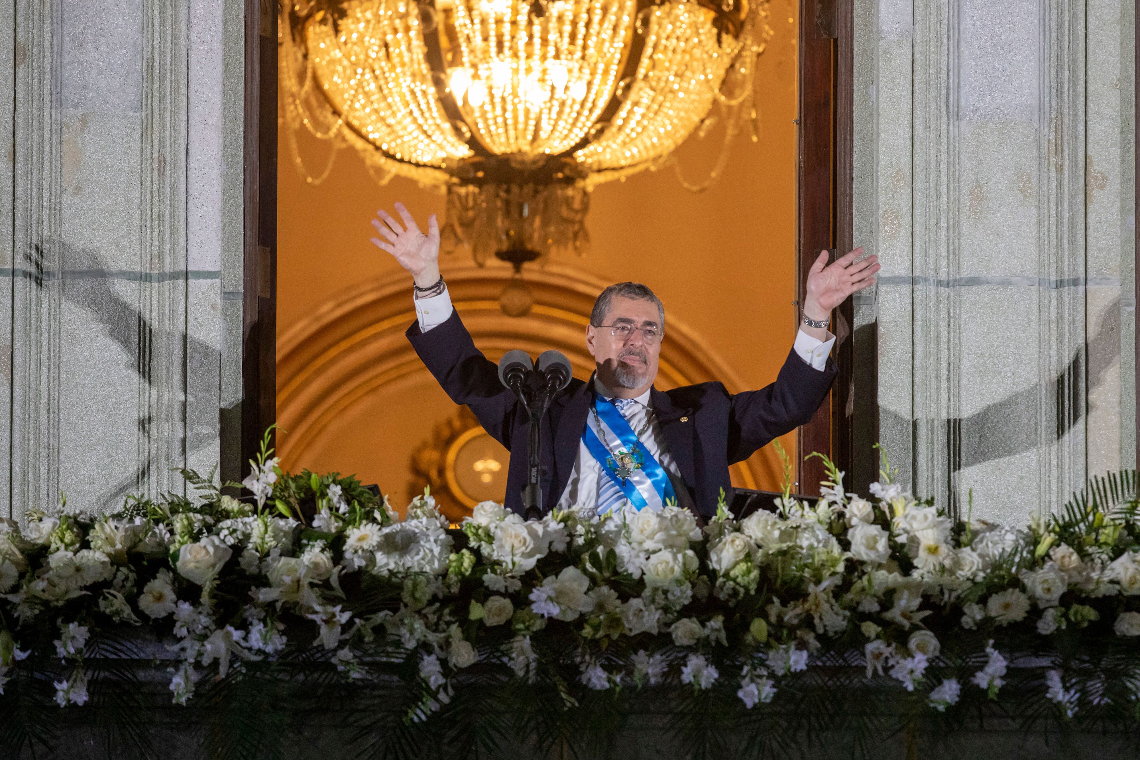 Guatemalan President Bernardo Arévalo raises both arms in the air while outside a palace after being sworn in. It is nighttime. There are rows of white flowers in front of him. 
