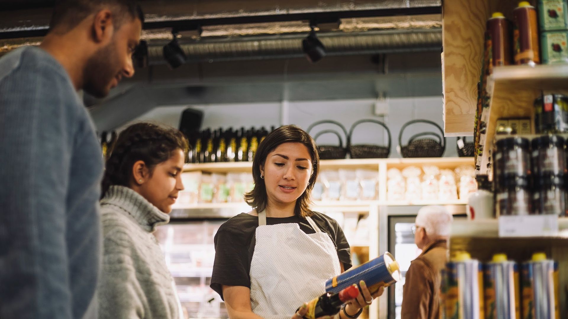 A woman in a white apron shows a bottle to two customers in a shop; shelves behind her hold beverages and snacks under warm lighting.
