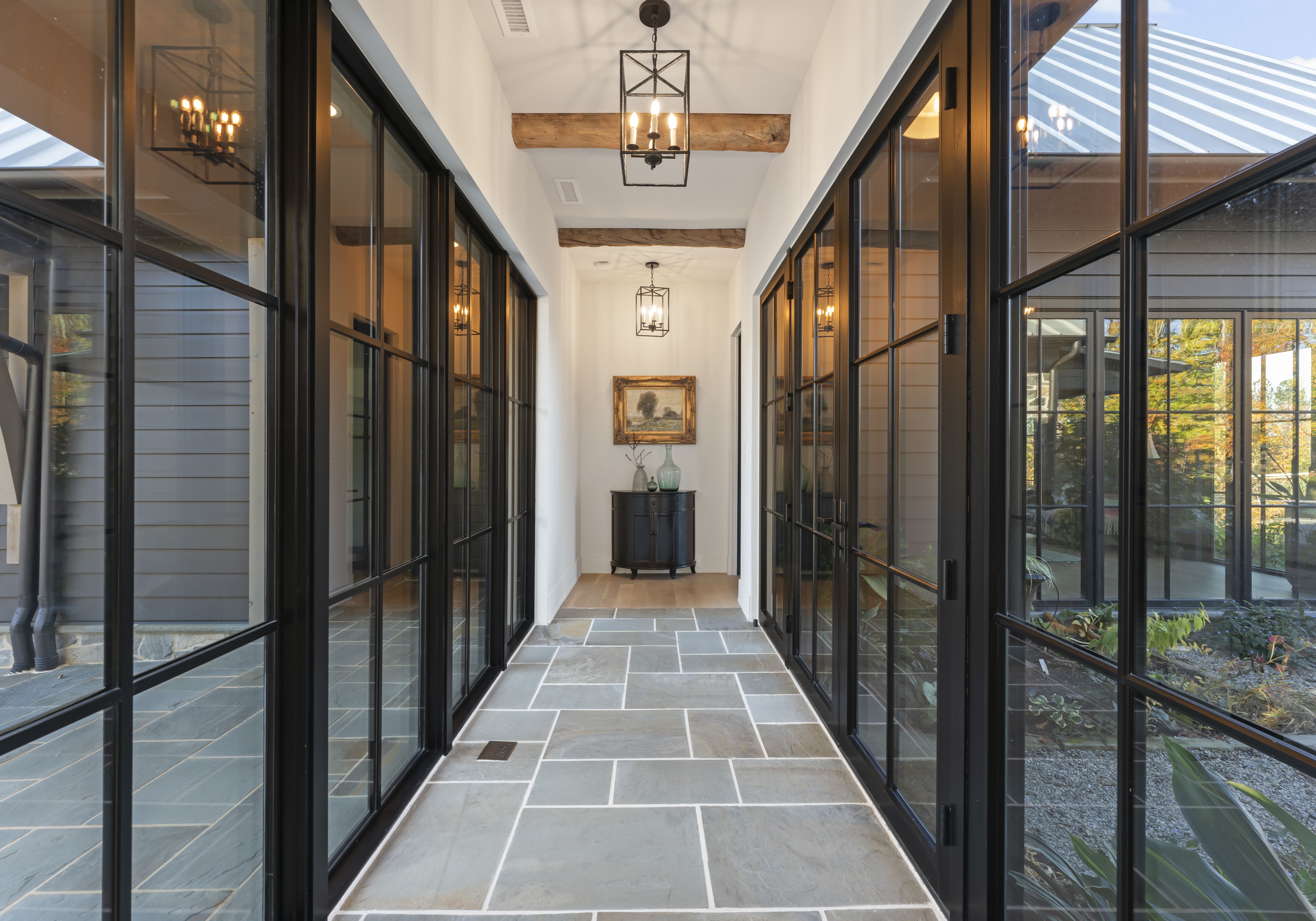 Bright glass-walled corridor with black metal frames, gray slate tile floor, and exposed wooden ceiling beams. At the far end, a dark round console with vases under a framed painting; warm pendant lights glow.