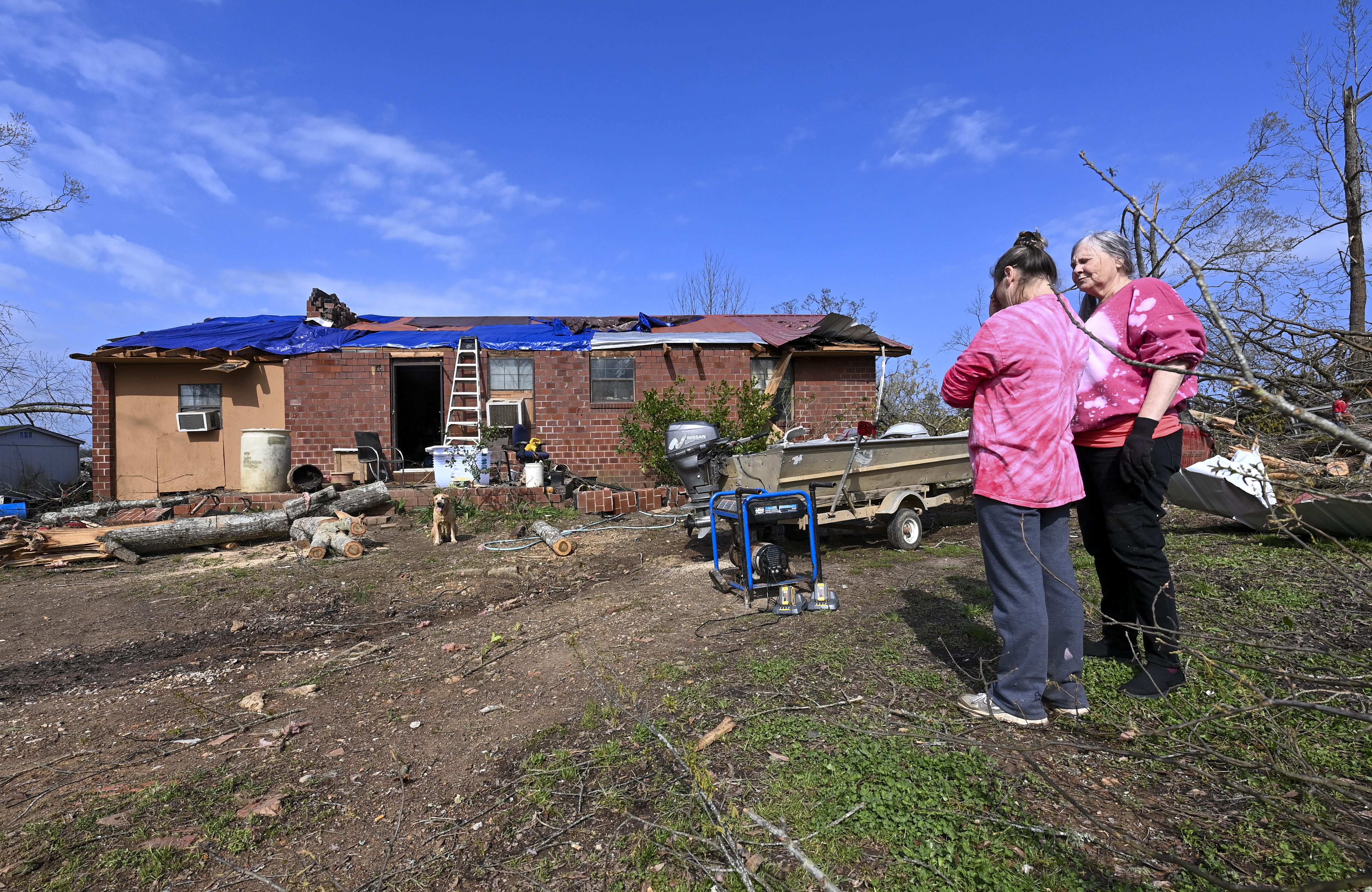 Photo of two people standing outside a destroyed home, one comforting the other