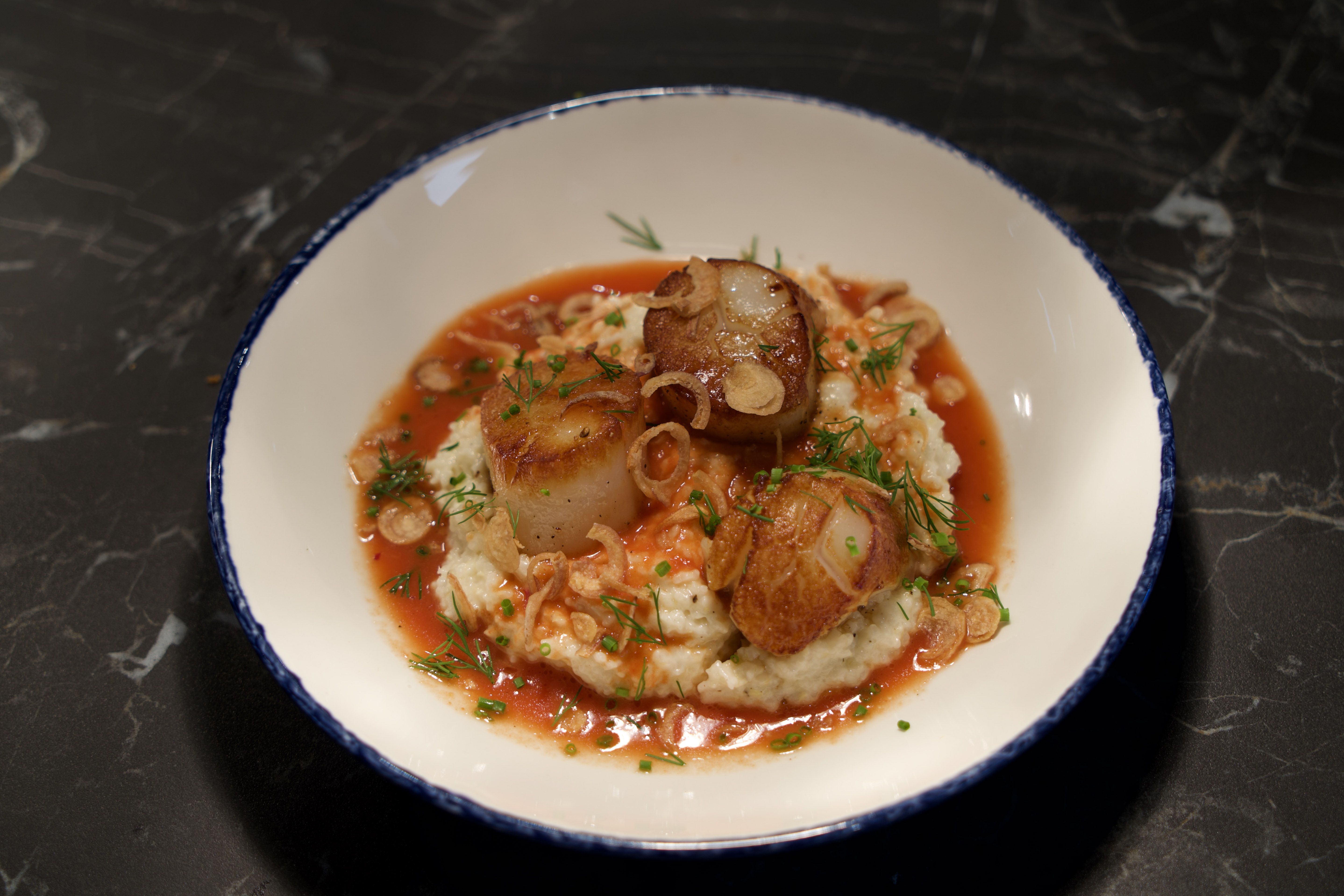 Seared scallops on creamy white risotto, garnished with green herbs, thin crispy shallots, and a red sauce in a white bowl with a blue rim on a dark marble surface.