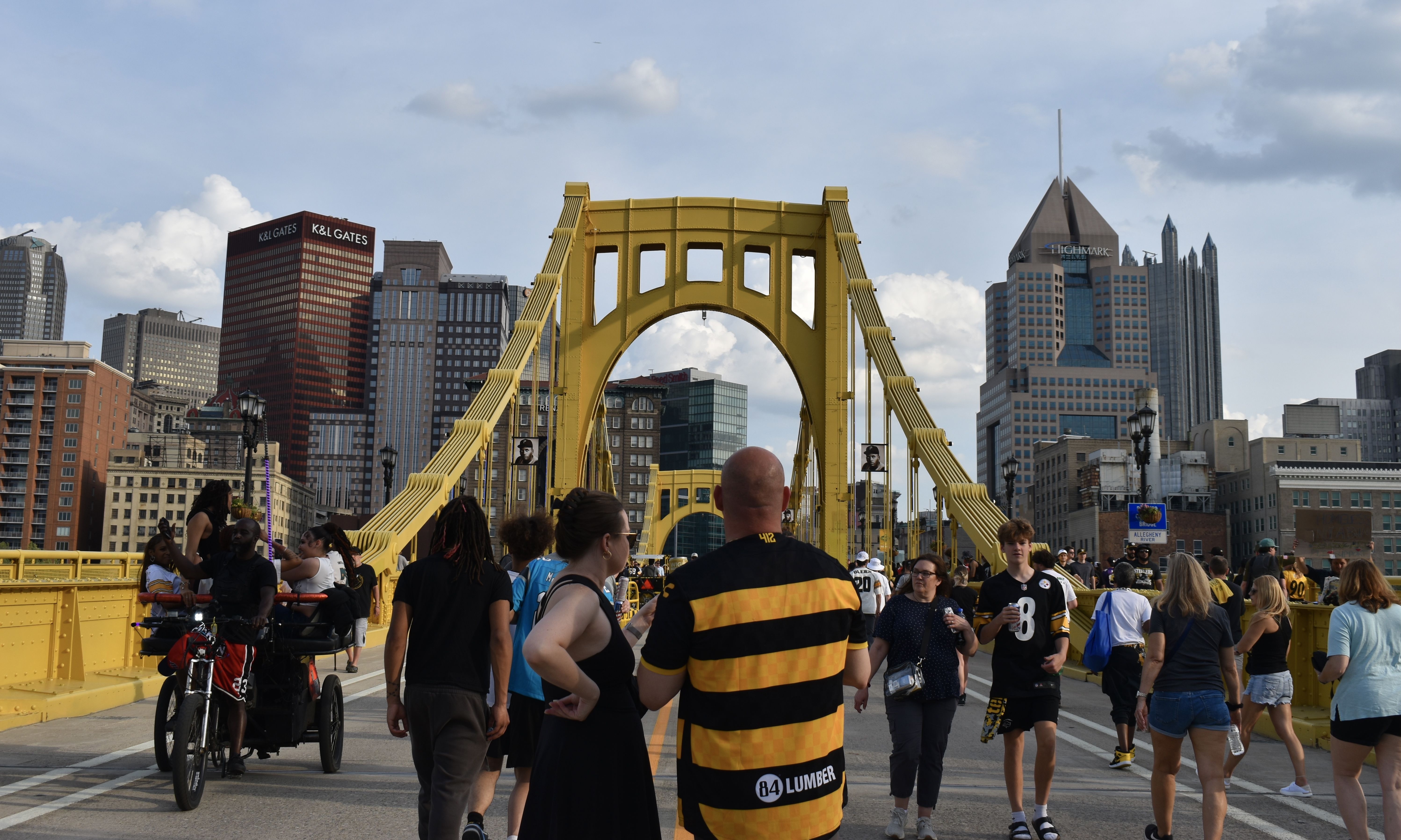 Busy pedestrian bridge in bright yellow, with people walking toward the camera; a city skyline of tall office buildings rises in the background under a blue sky.