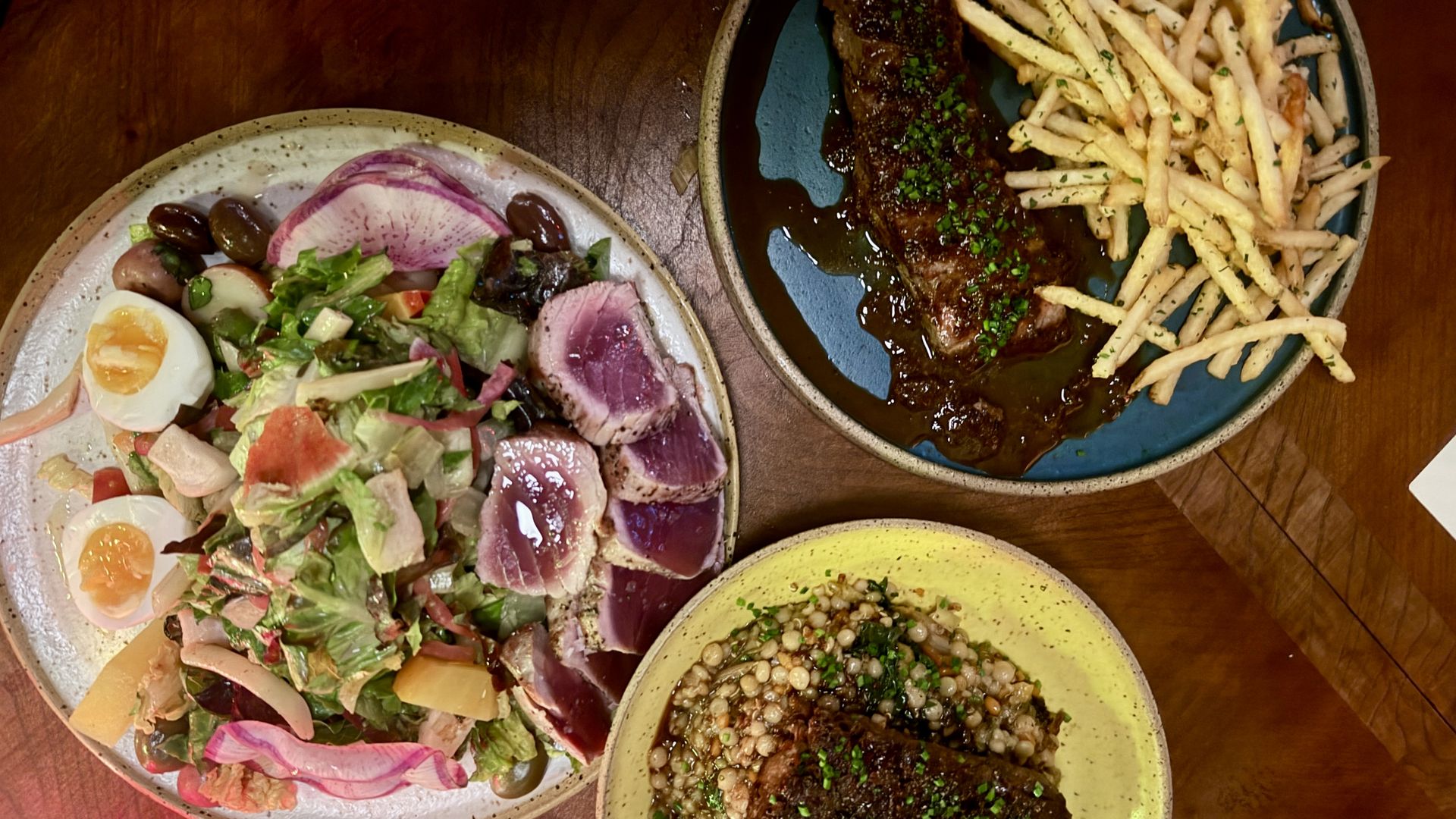 Top-down view of a meal on three plates: a beef steak in brown sauce with fries and herb garnish; a colorful salad with radishes, olives, greens, and a boiled egg; and a grain-lentil dish.
