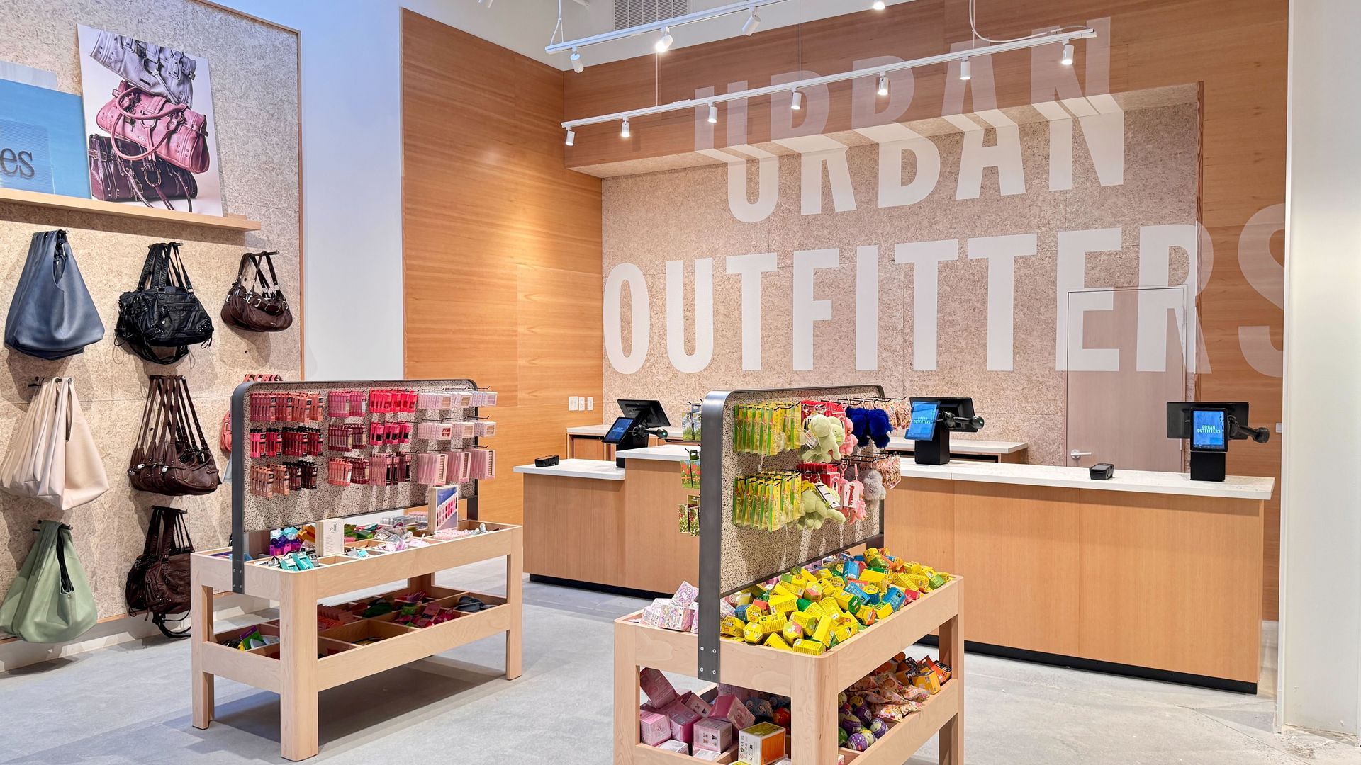 Bright store interior with wooden fixtures, handbags on the left, tables of colorful goods, and a large wall graphic reading "ORDAN OUTFITTERS" in white letters.