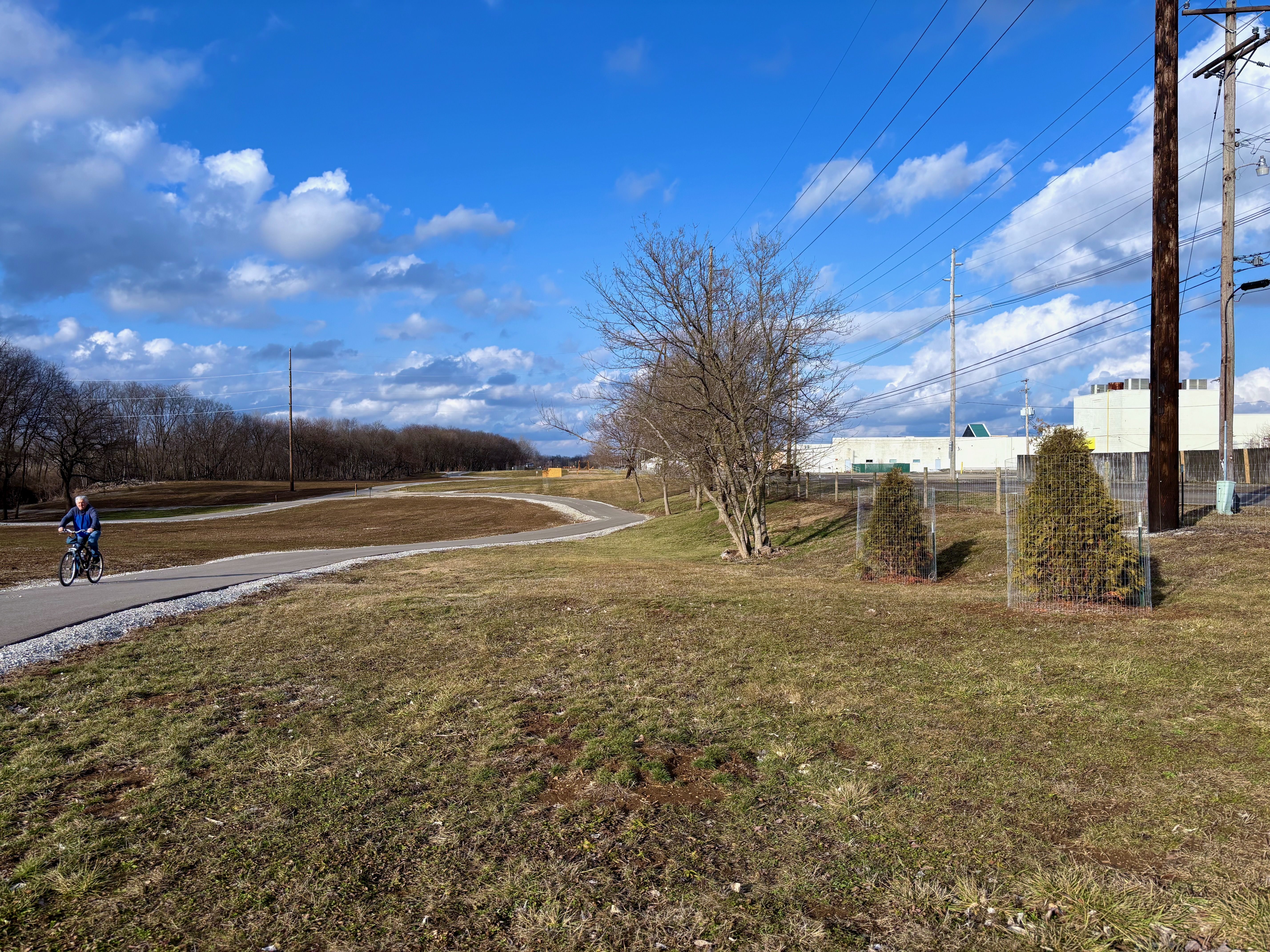 A person rides a bike on a paved winding path through a grassy area with leafless trees under a partly cloudy blue sky, with utility poles and a white building in the background.