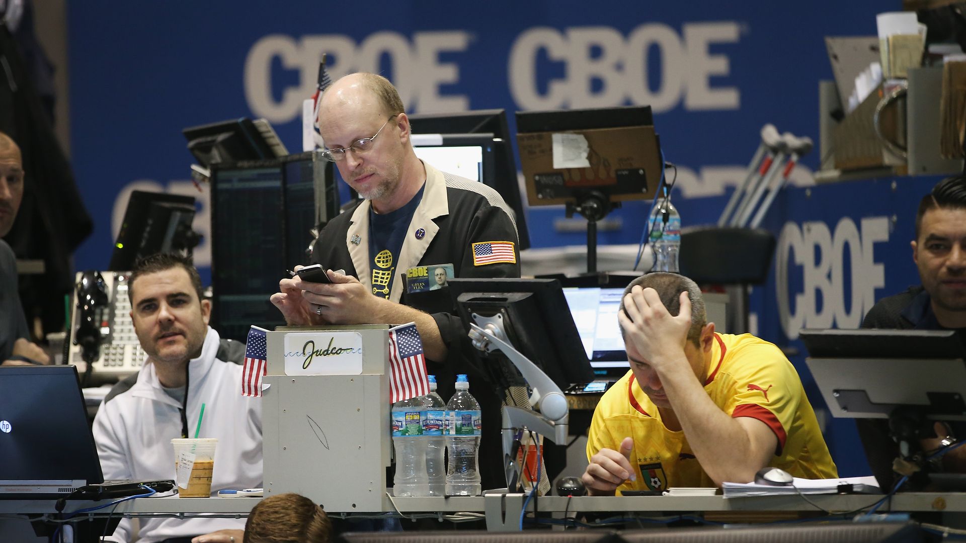 In this image, two men sit behind computers on a trading floor and one man stands between them, looking at his phone.