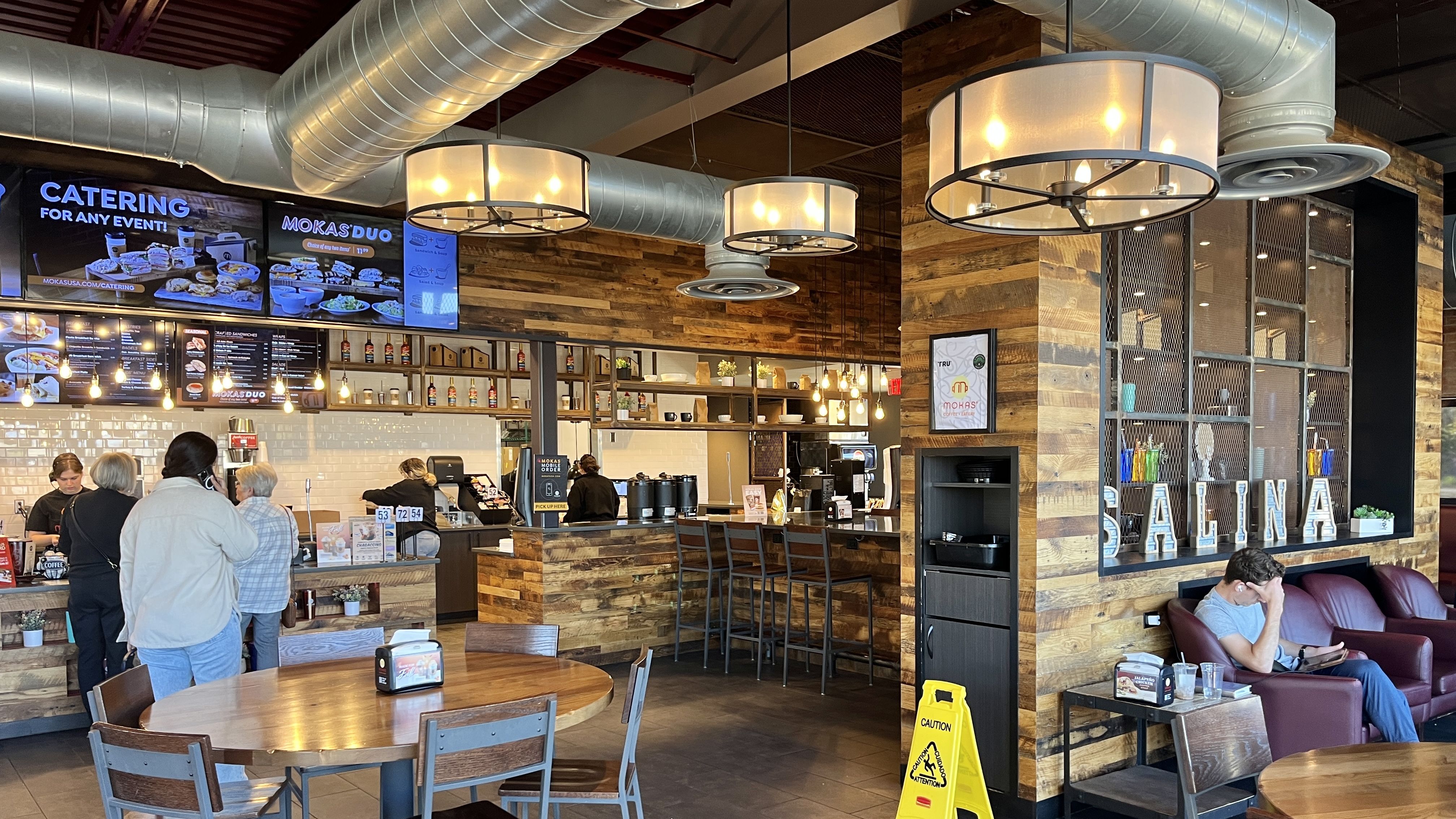 Interior of a cozy café with wooden walls and round hanging lights, customers ordering at counter with menu boards, and a person seated on a red couch near large window with letters spelling SALINA.