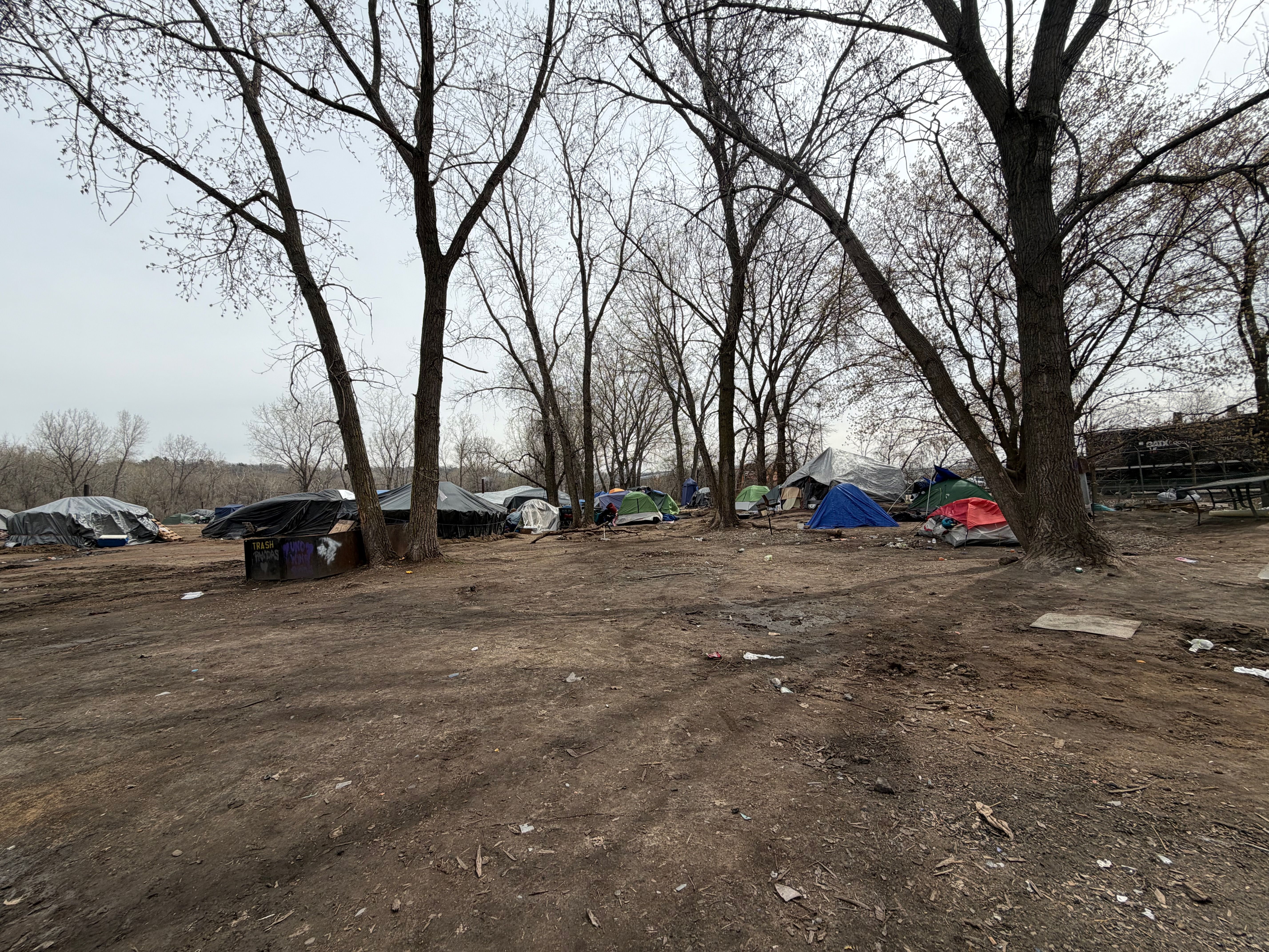 A row of leafless trees over a dirt clearing, with colorful tents pitched beneath. Trash and litter scattered on the ground, a bleak overcast sky above.