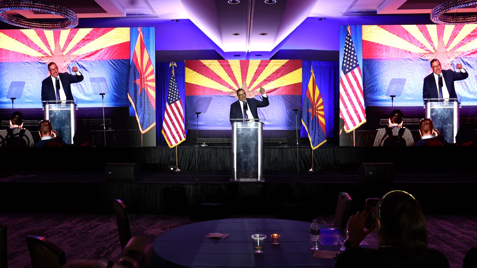 A man in a suit speaking passionately at a podium with raised fist, flanked by large Arizona and U.S. flags with an audience and photographers in a dimly lit room.