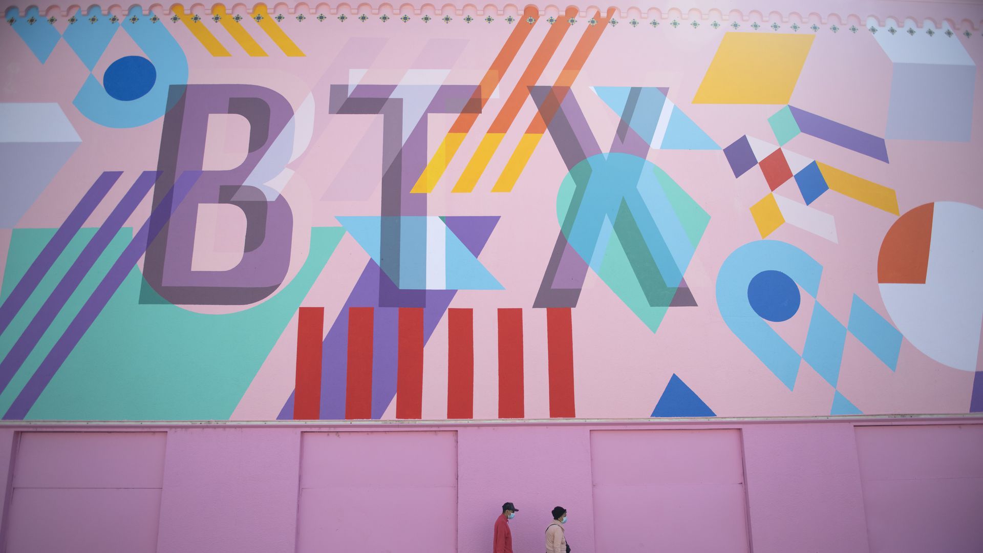 A large mural with the letters BTW in pastel colors is the backdrop as two people with medical masks walk by 