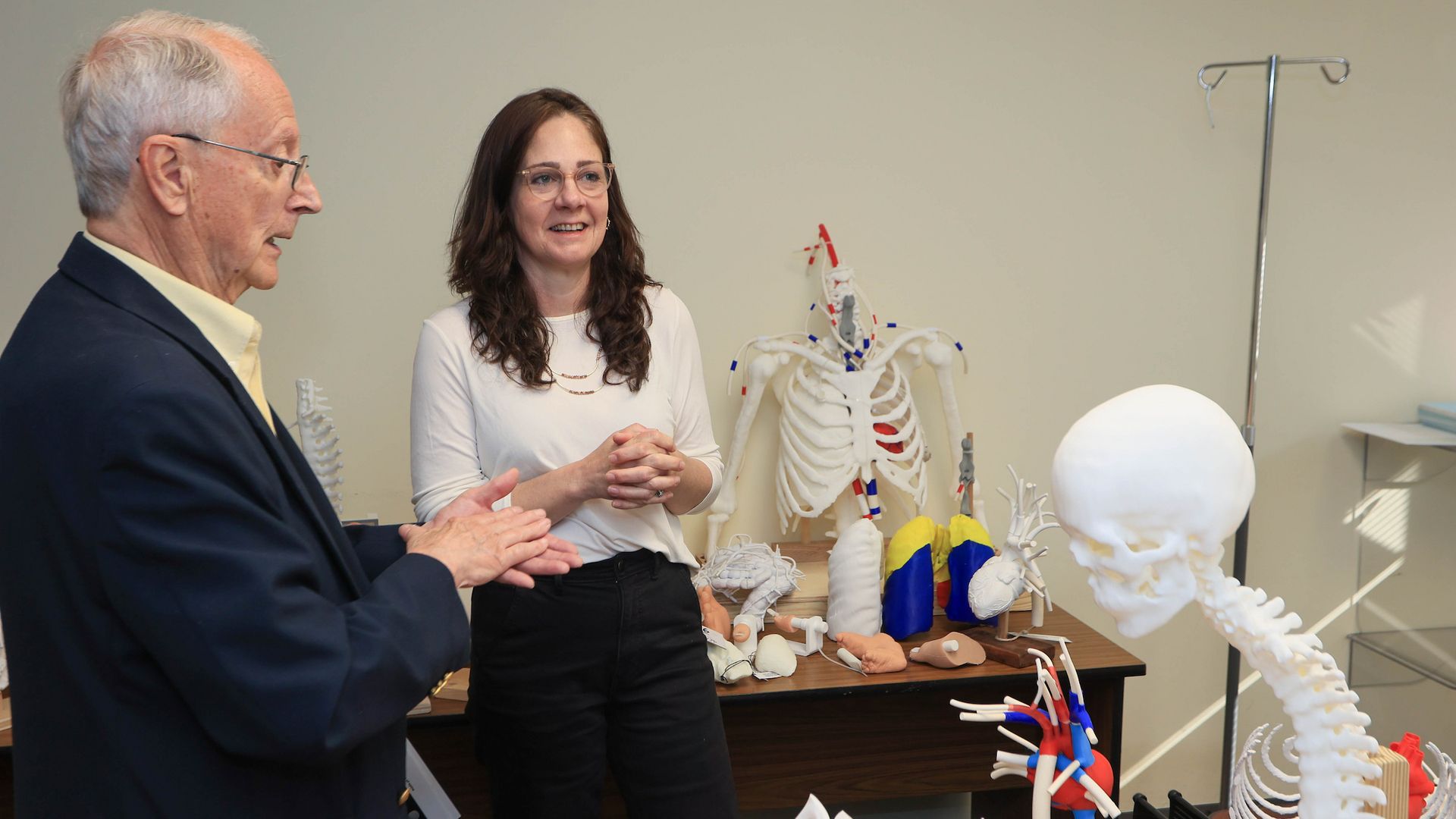 Two adults in a classroom, an older man in a dark suit and a younger woman with glasses, talking beside a table full of anatomical skeletons and medical models.