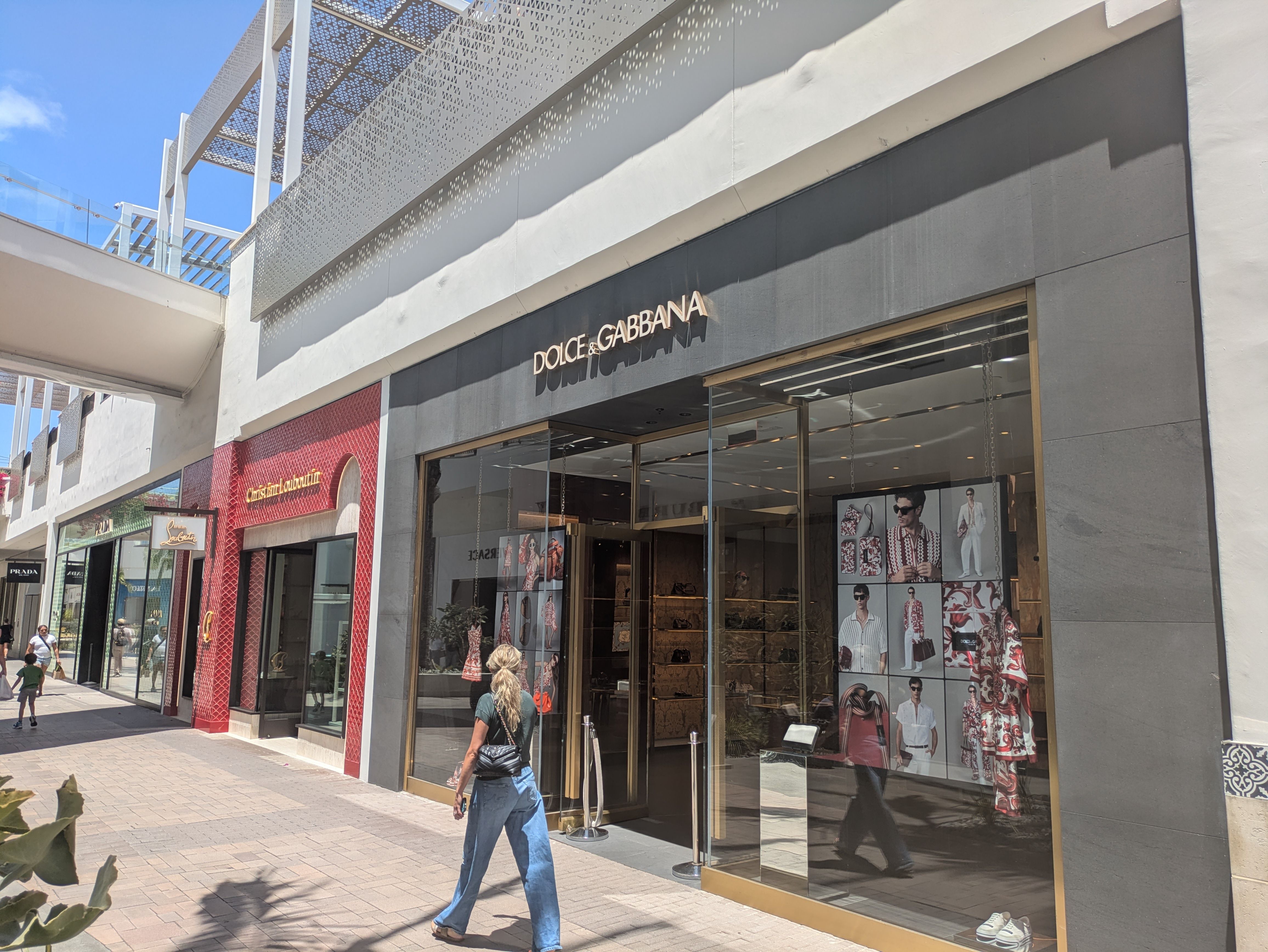Dolce & Gabbana storefront on a sunny street with glass display windows, fashion posters, and a gray facade. A perforated metal awning overhead casts shadows as pedestrians walk by.
