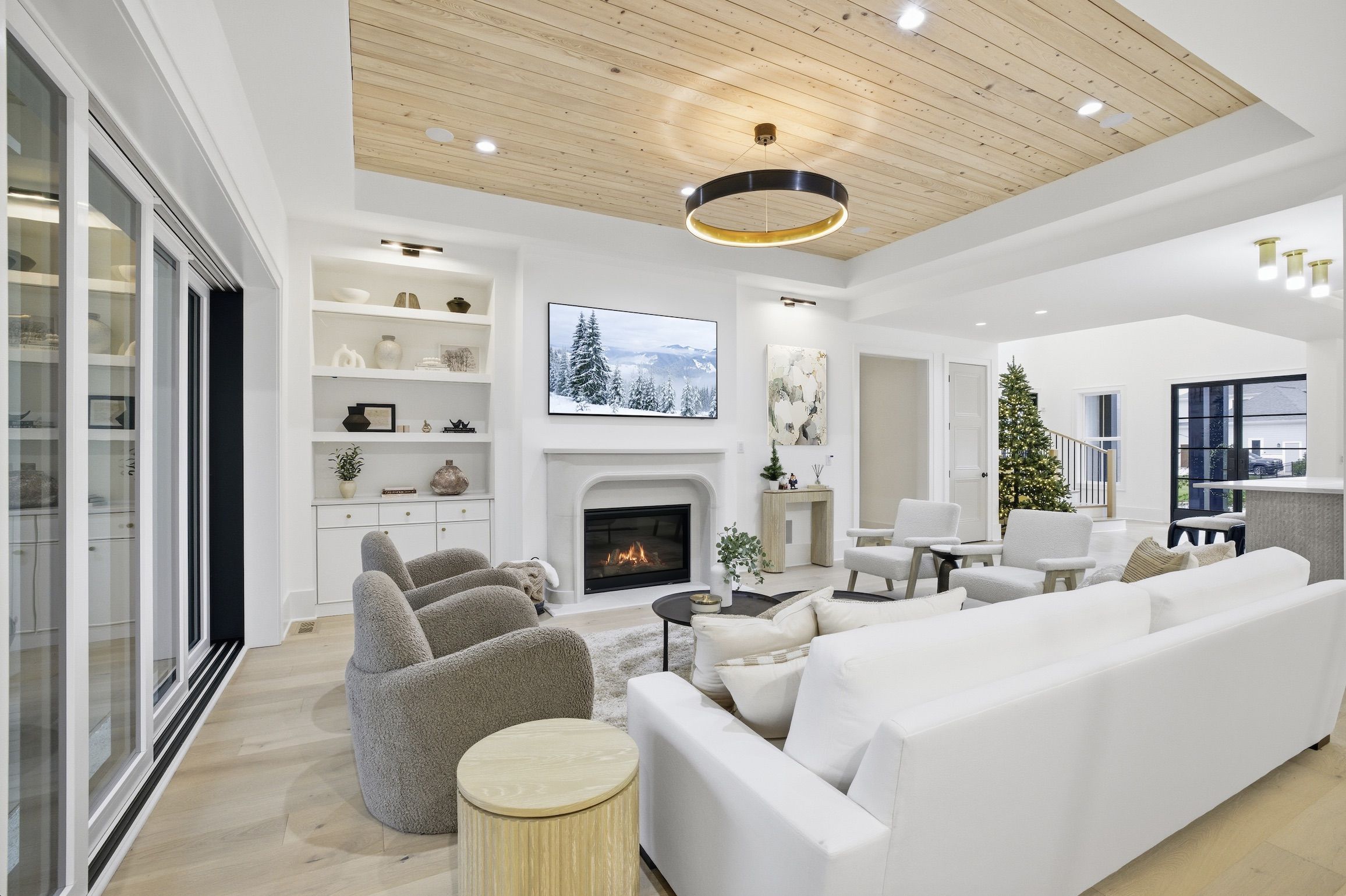Bright living room with white walls, wood ceiling, beige chairs, white sofa, fireplace, TV showing snowy forest, Christmas tree, and modern decor with light hardwood floors.