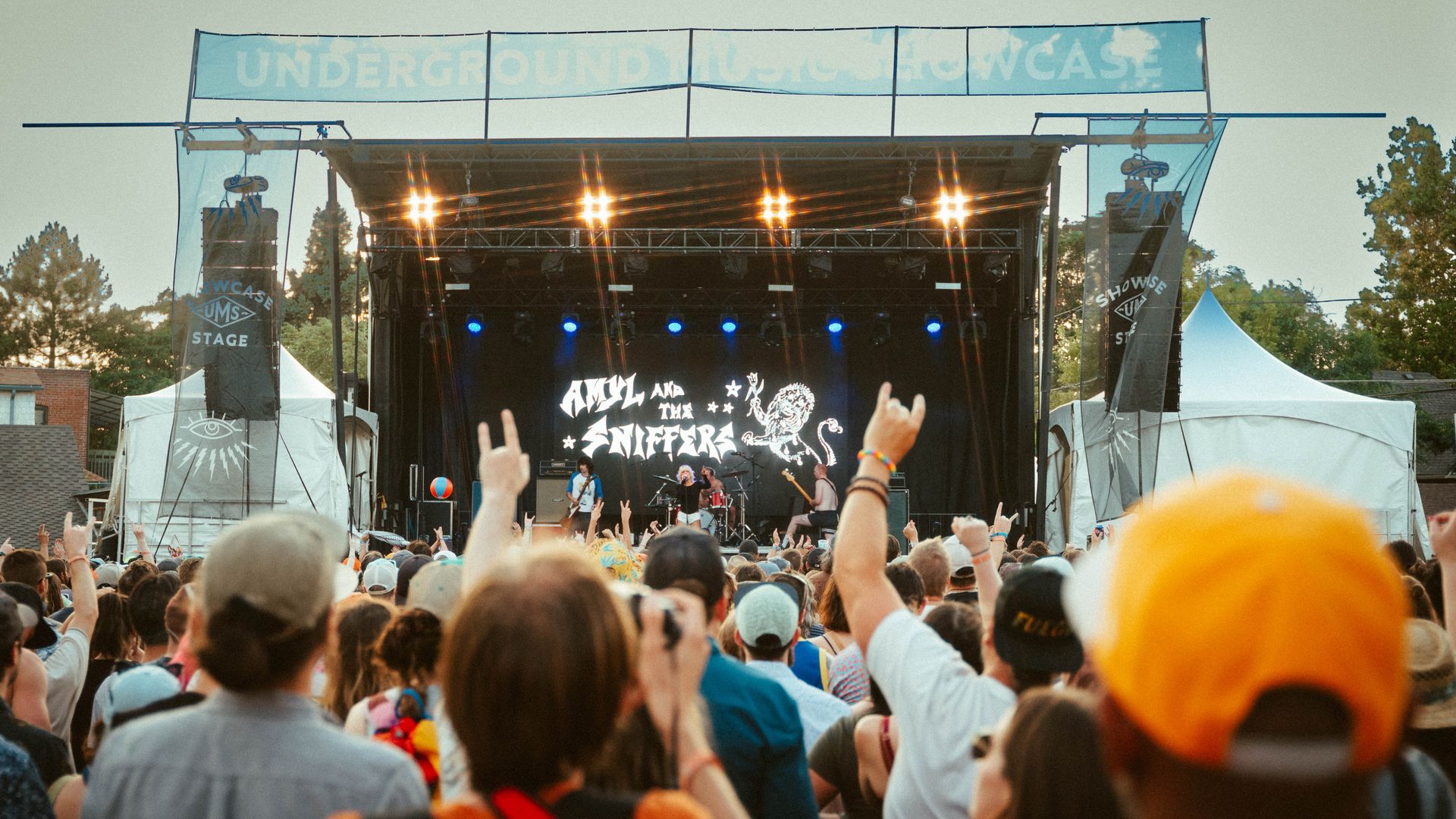 Outdoor concert stage with bright lights, a band performing, and a cheering crowd with hands raised. Large blue banners above the stage, trees in the background.