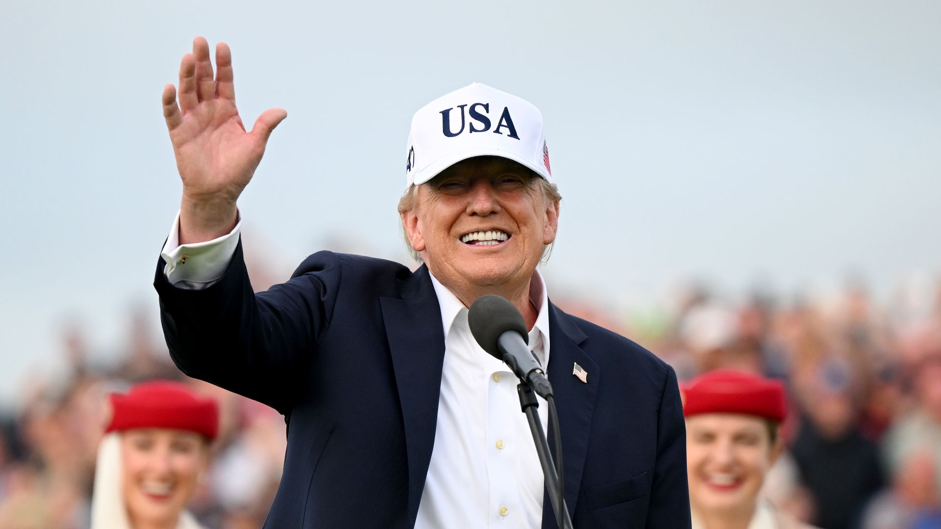 Close-up of President Donald Trump wearing a white cap marked "USA", waving to a cheering crowd from a podium with microphones; a blurred audience behind him.