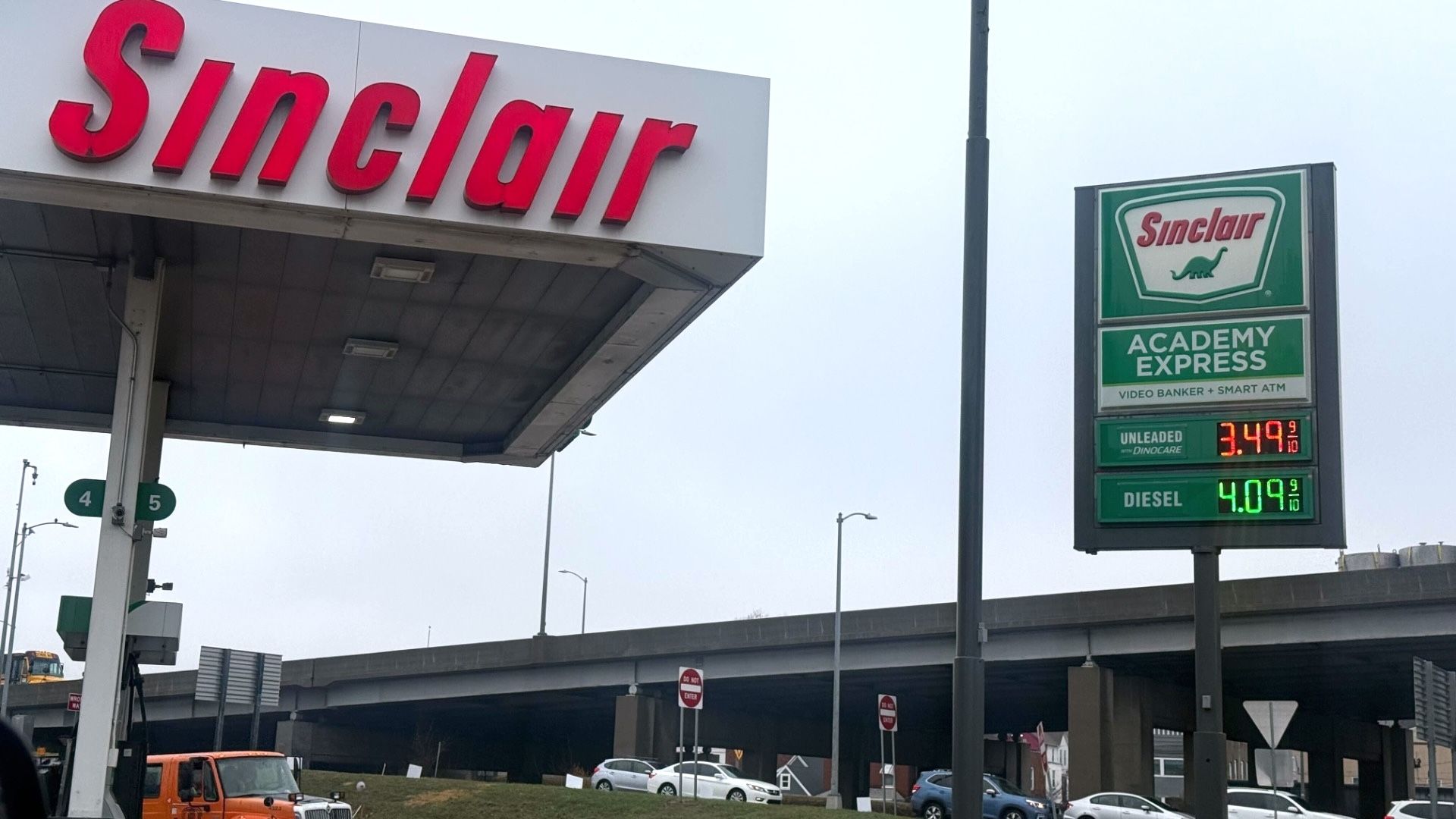Sinclair gas station with red signage and green price board showing unleaded gas at $3.49 and diesel at $4.09, located near a highway overpass on a cloudy day.