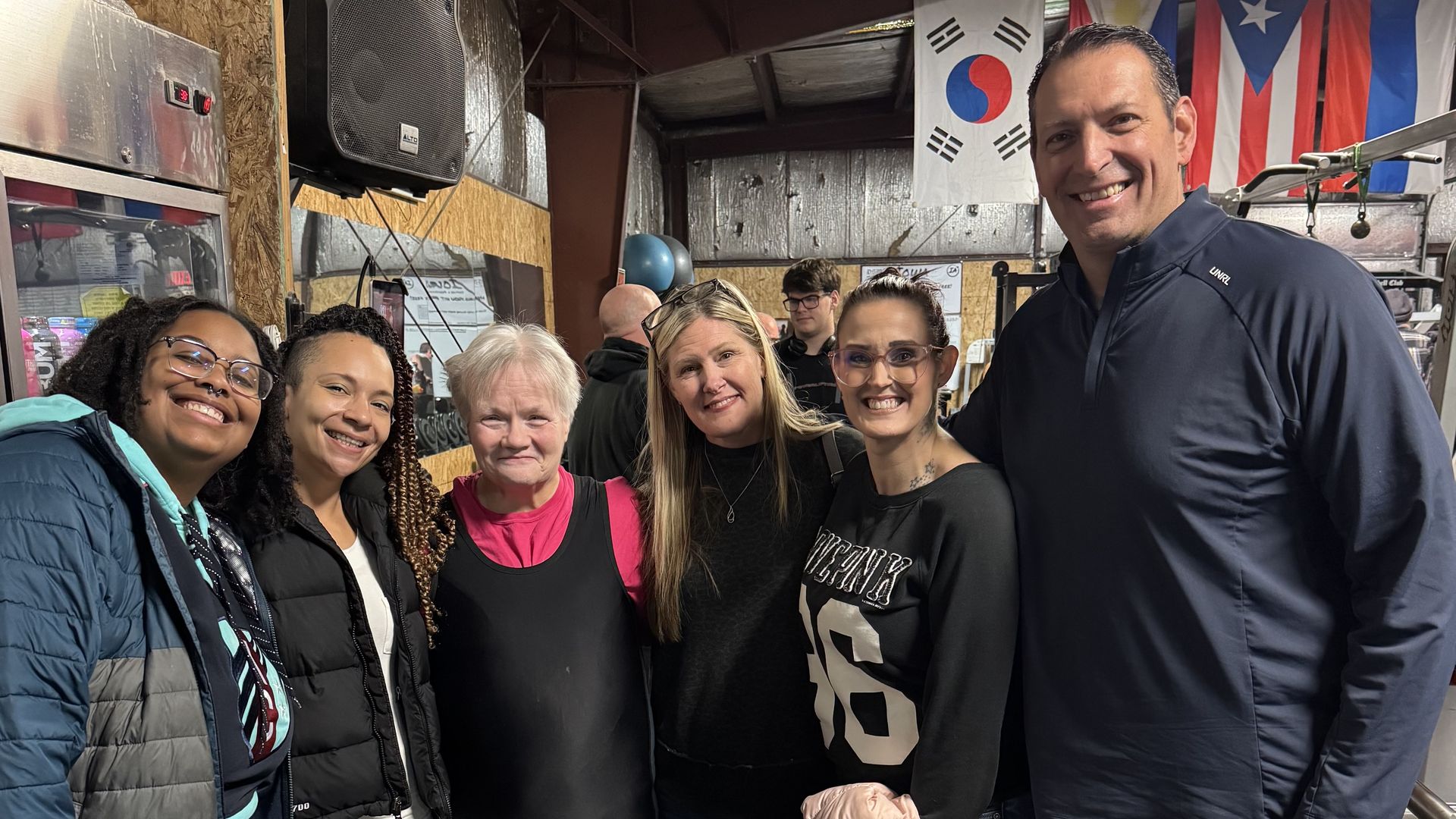Seven adults pose for a group photo in a gym, with flags on the wall including the South Korea and United States flags. They smile and wear athletic jackets and workout clothes.