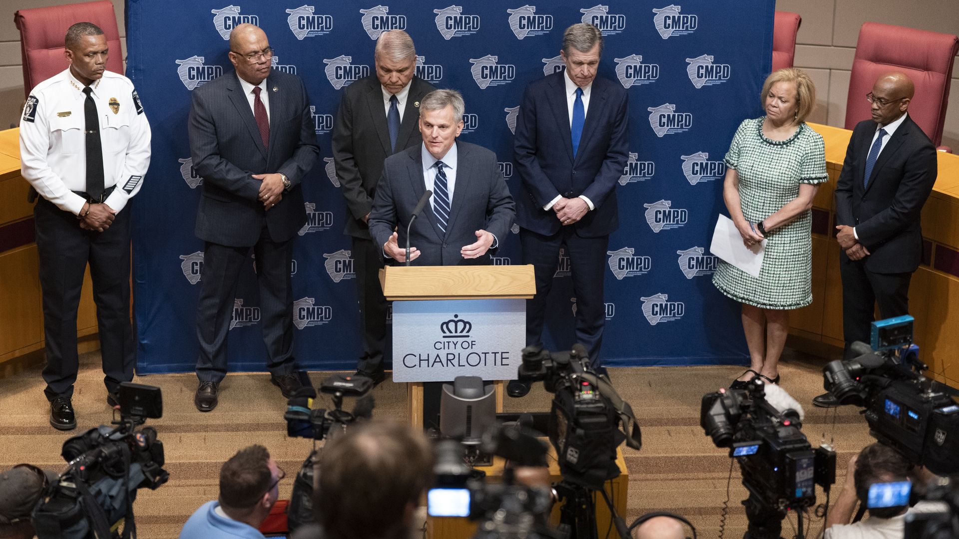 Press conference at City of Charlotte as a man in a suit speaks at a wooden podium; CMPD backdrop with logos; six officials and a police officer stand behind him, cameras in the foreground.