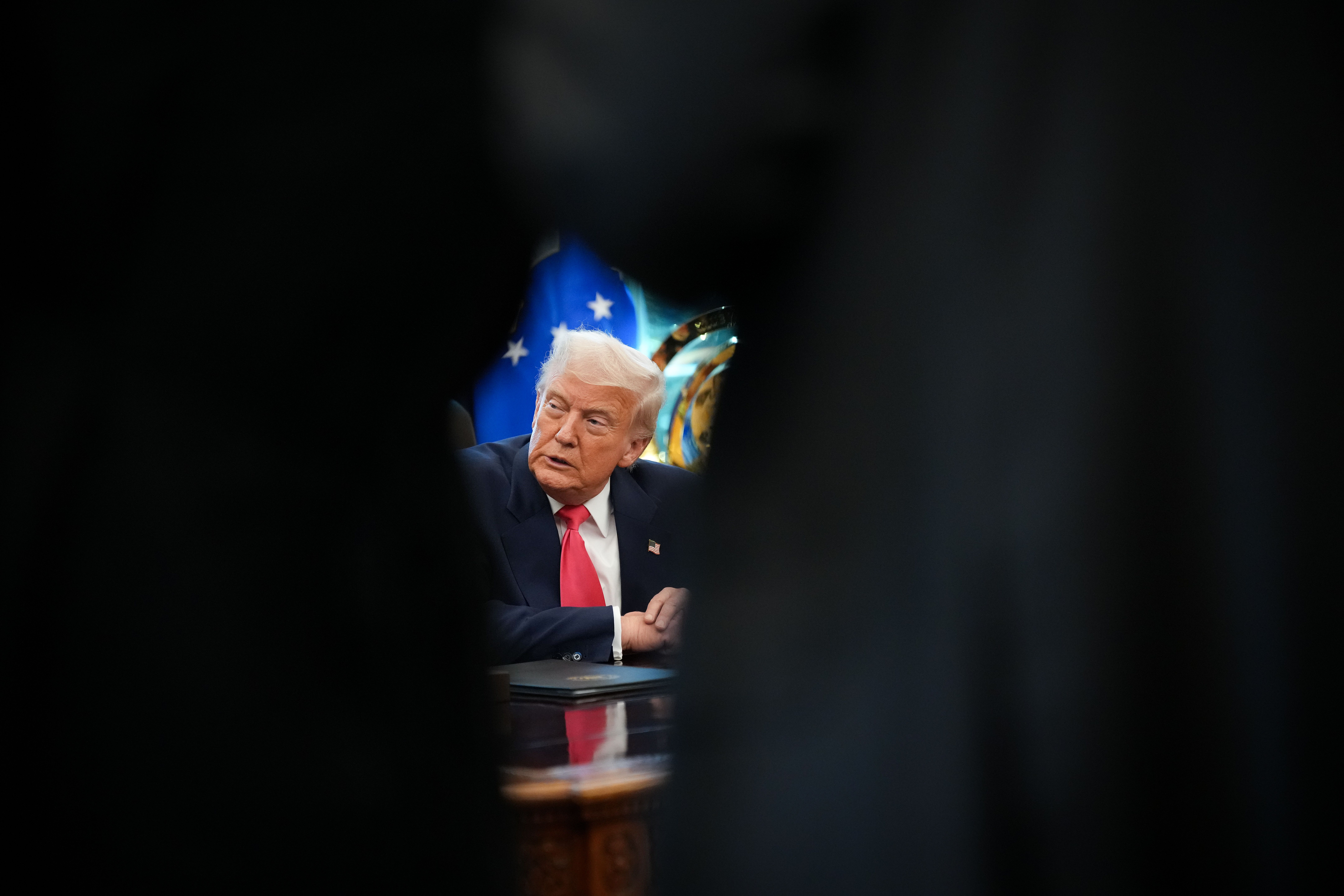 Former President Donald Trump in a navy suit and red tie, sitting at a desk, seen through a narrow gap with a blurred blue background featuring white stars.
