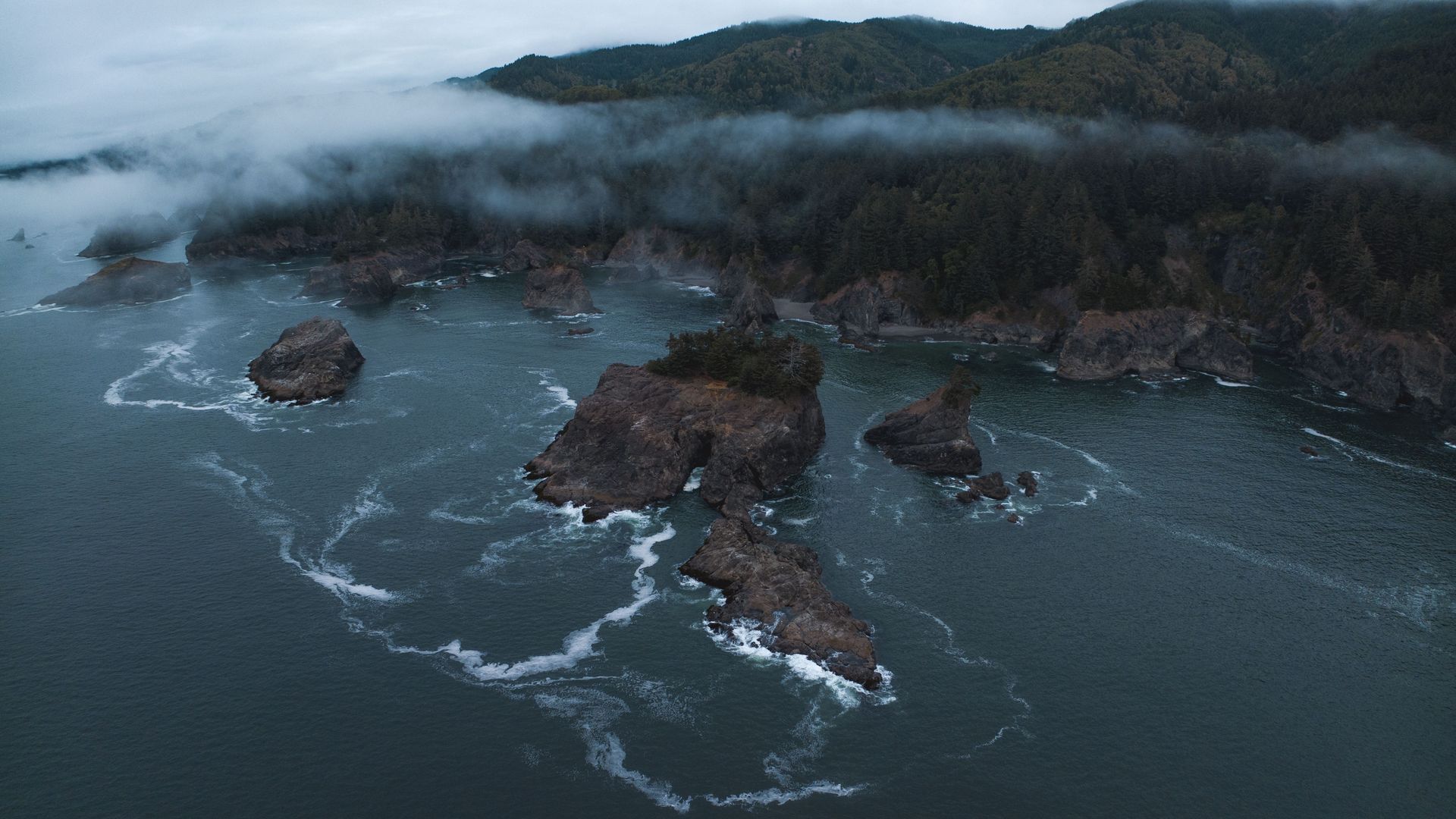 Aerial view of rocky islands with trees in dark blue ocean waters, white waves swirling around, and mist hovering over forested green hills in overcast weather.