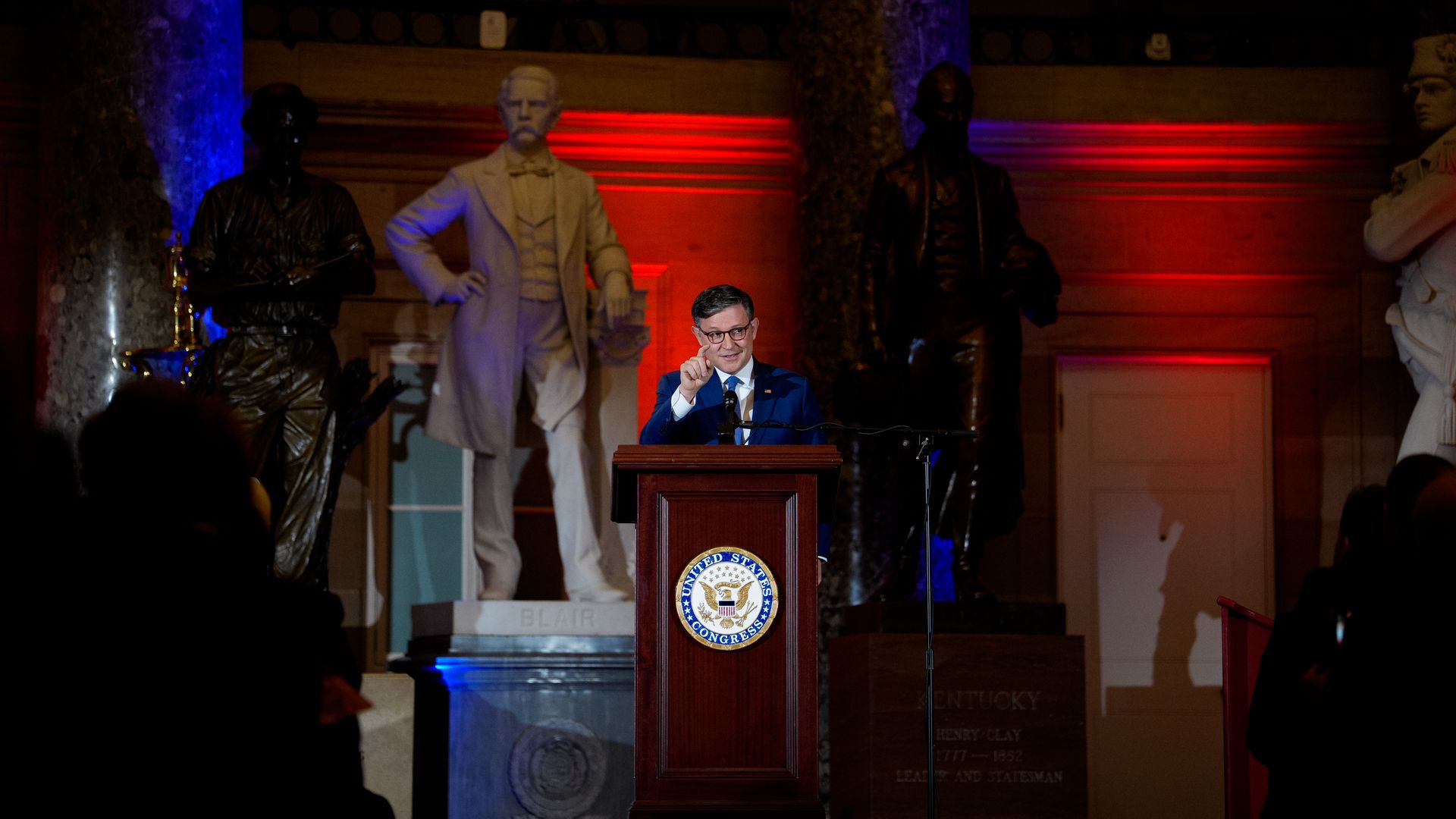 A man in a blue suit speaks at a wooden podium with the United States Congress seal, flanked by statues in a museum-like hall under blue and red lighting.
